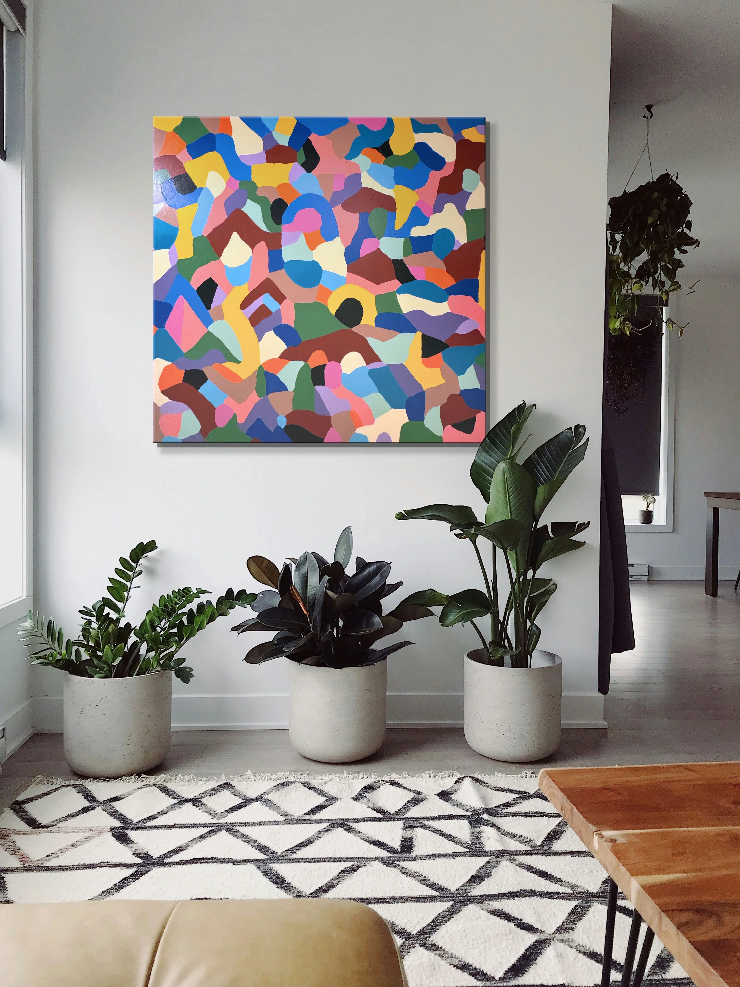 A colorful abstract painting hanging on a wall in a living space with house plants and a rug.