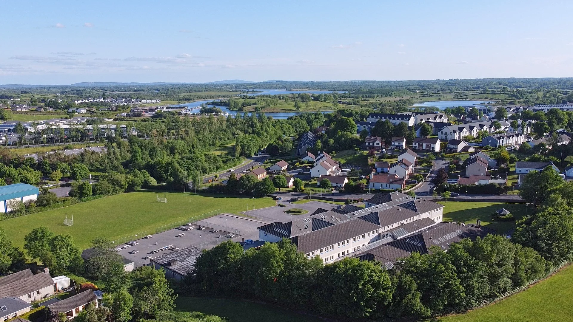 Aerial view of a suburban area with green fields, trees, a school with a parking lot, and residential houses near a river under a partly cloudy sky.