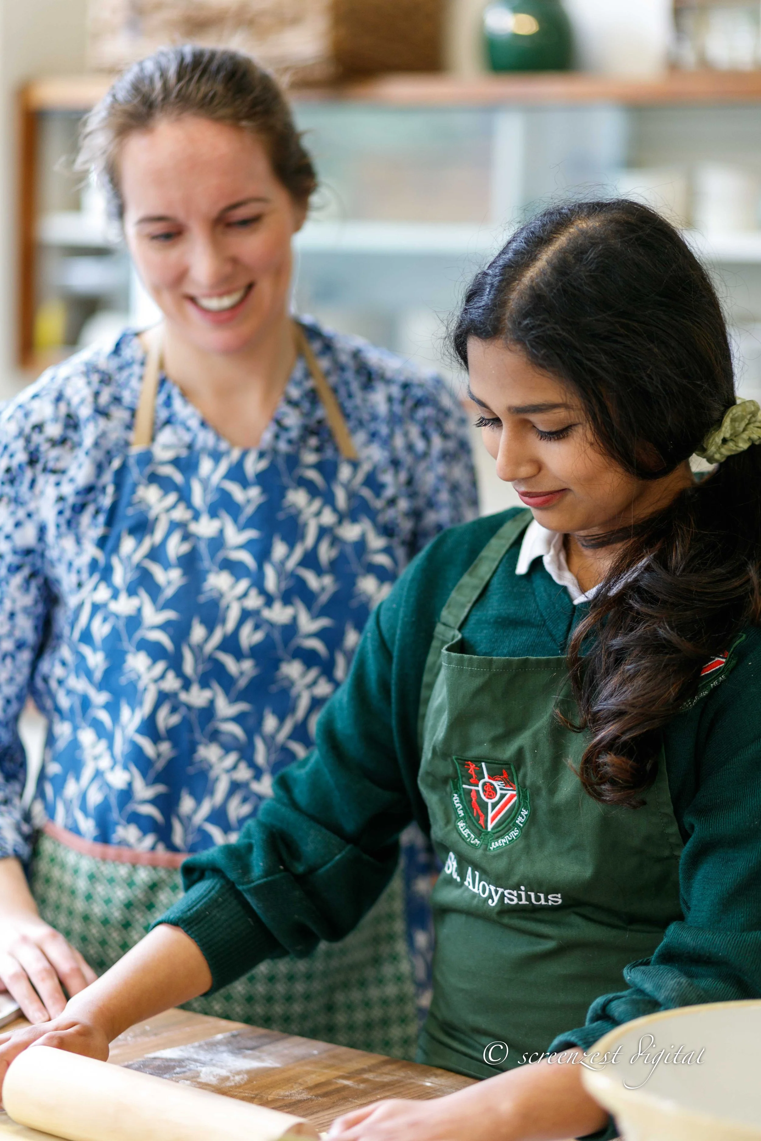 A young girl rolls out dough with a wooden rolling pin while a woman watches and smiles in a kitchen.