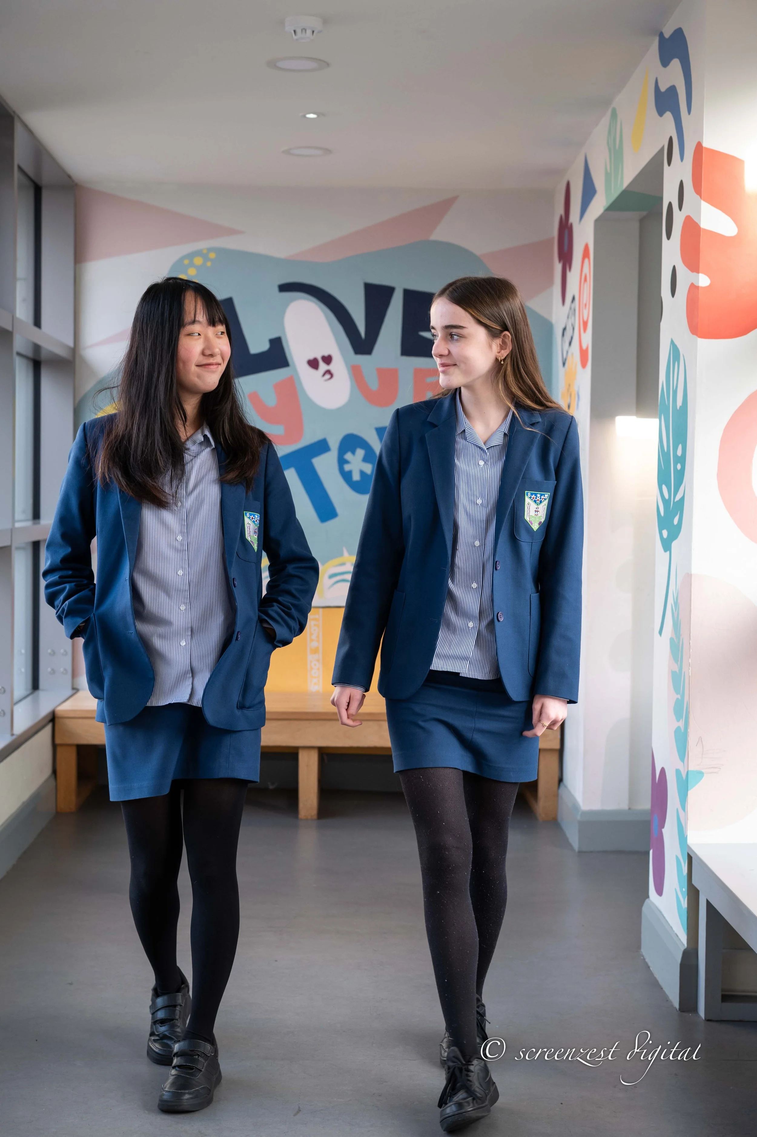 Two schoolgirls in navy blue blazers and skirts walking together in a hallway with colorful wall art that says "LOVE" and "“You."