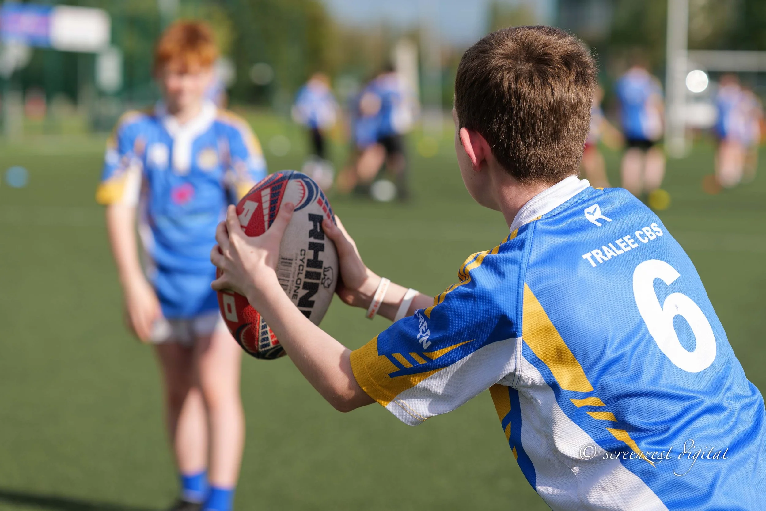 A young rugby player in a blue and yellow jersey prepares to pass a rugby ball during a game on a grassy field, with other players in similar uniforms blurred in the background.