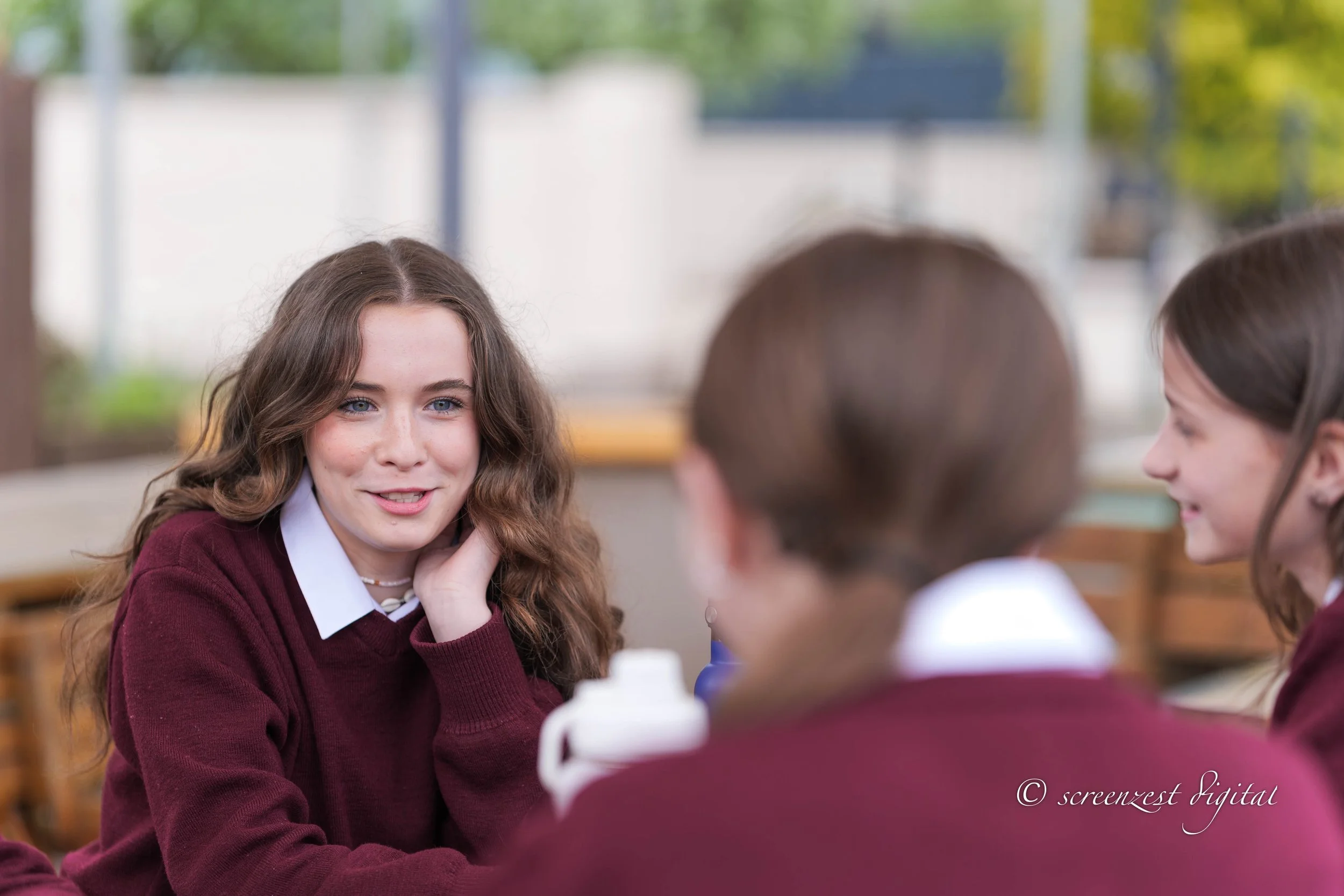 Three girls in school uniforms having a conversation outdoors, with the girl in the middle smiling and resting her chin on her hand.