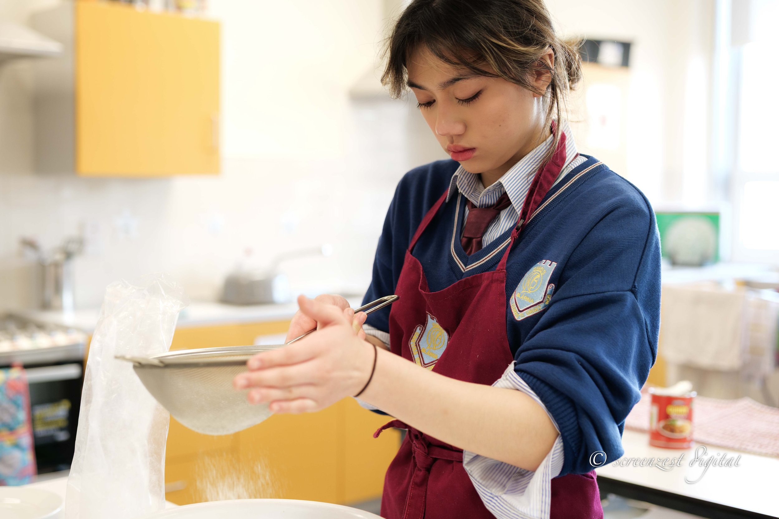 Young girl with short hair in a school uniform and apron, pouring flour from a plastic bag into a bowl in a kitchen.