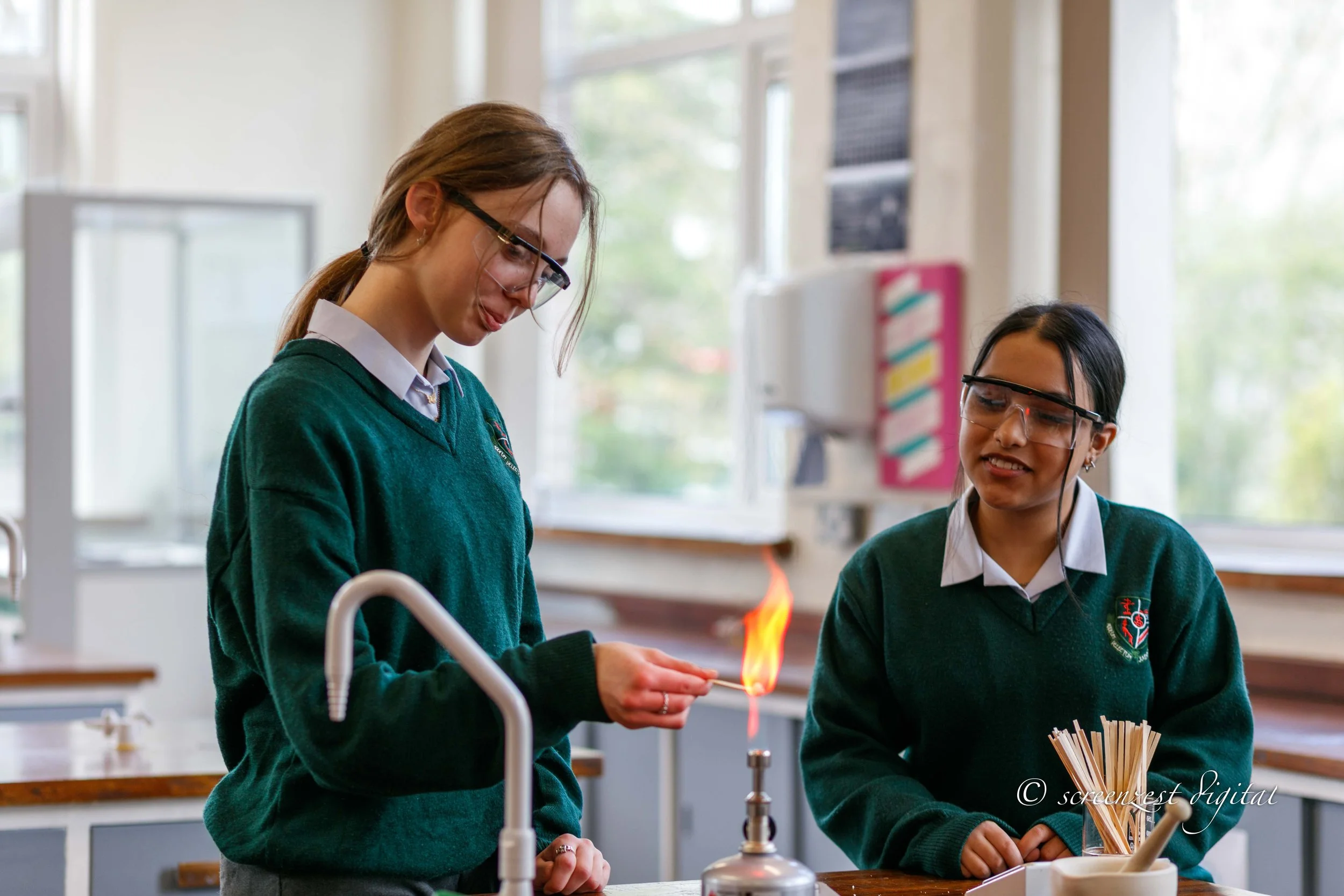 Two female students wearing safety glasses and school uniforms in a science classroom, conducting an experiment with a Bunsen burner, with one student observing a flame.