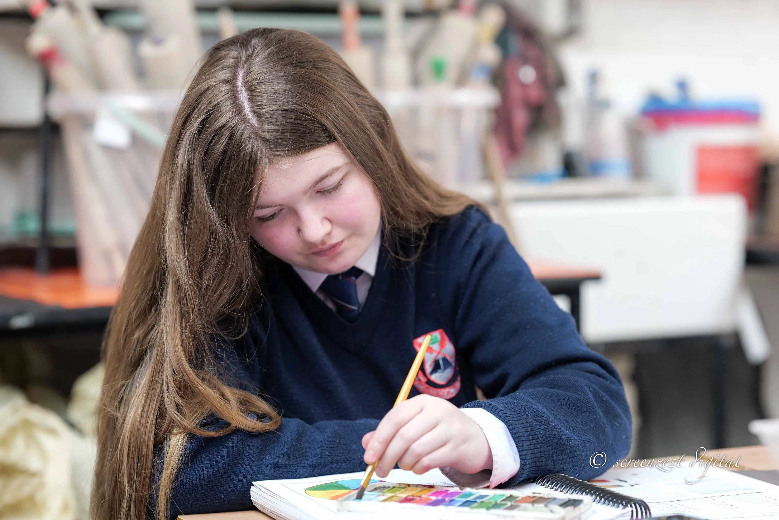 A young girl in a school uniform drawing or coloring on a notebook with a watercolor palette.