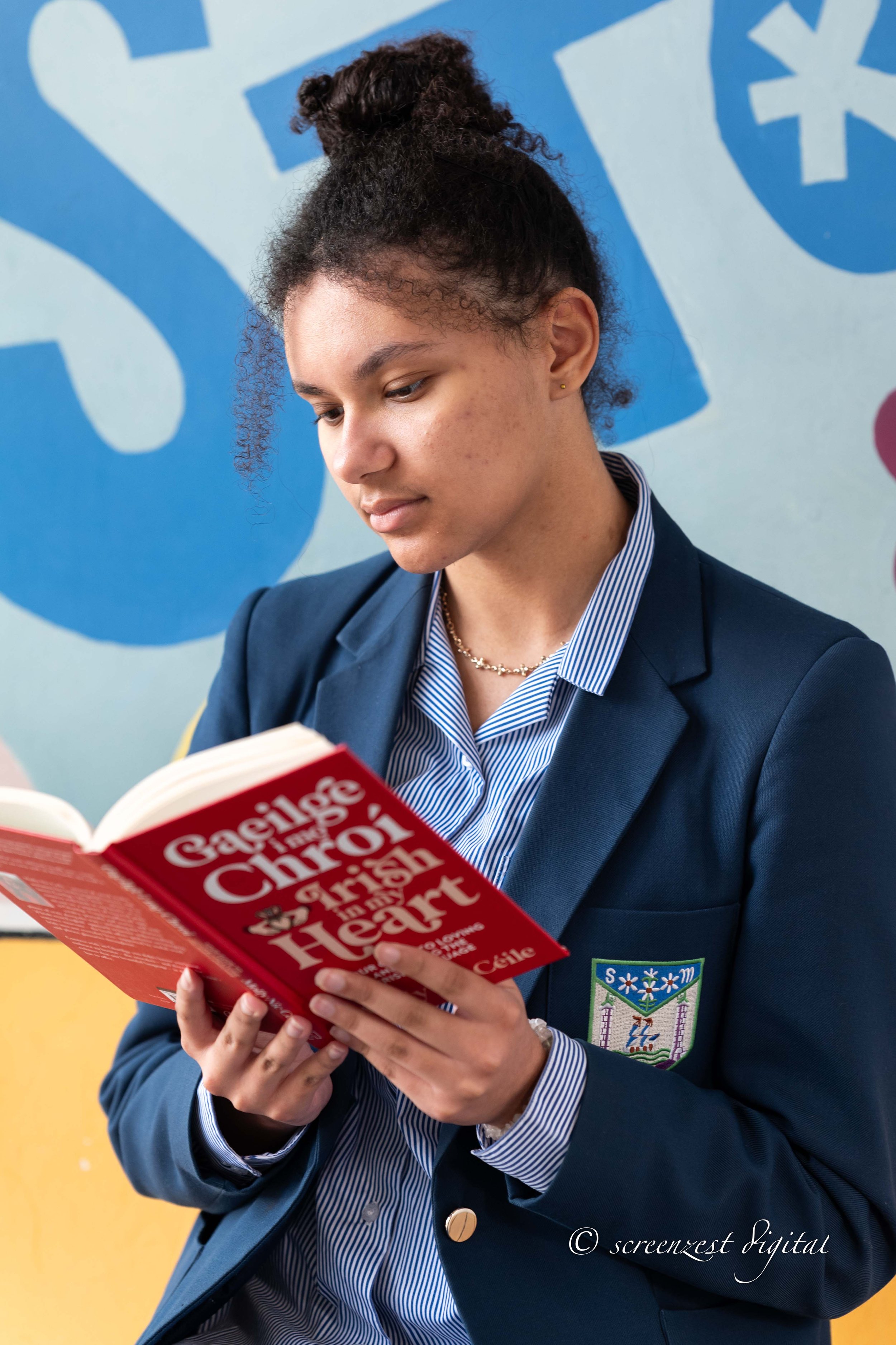 A young woman with curly hair tied up, wearing a blue blazer with a school emblem, reading a red book titled 'Gaeilge Timing Chroi & a High Heart' in front of a colorful wall.