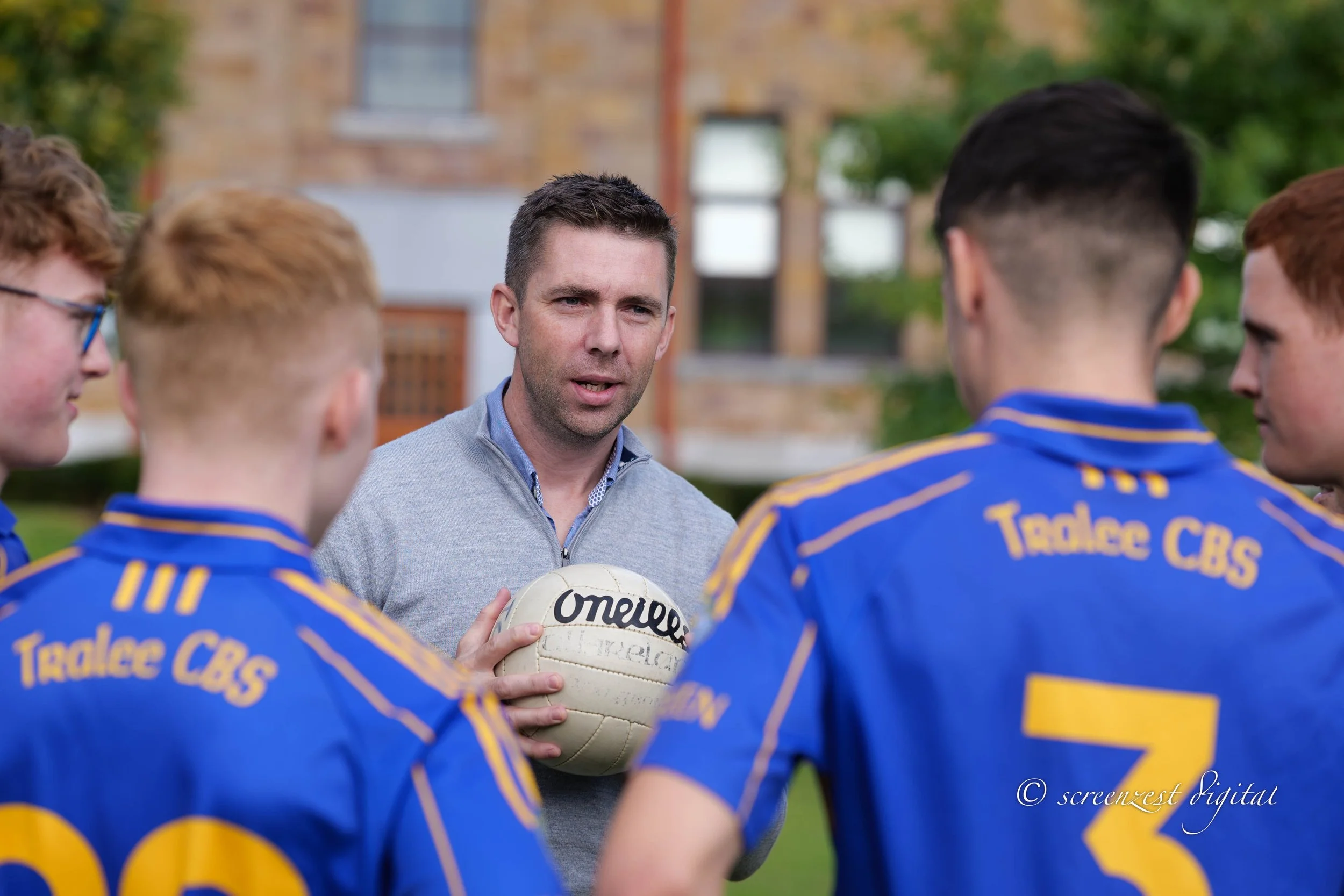 A coach or trainer giving instructions to a group of young soccer players outdoors, one holding a volleyball.