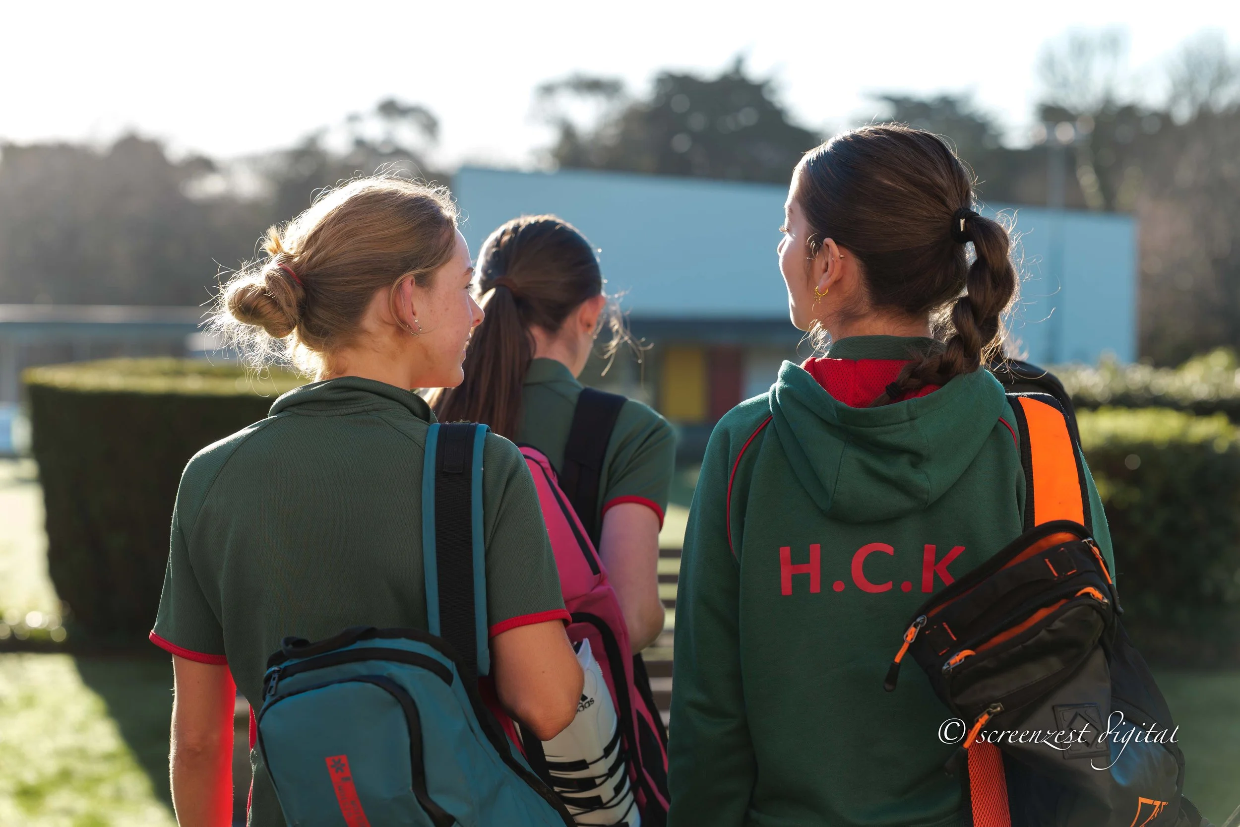 Three young women with backpacks standing outdoors, engaged in conversation, with a building and trees in the background during daytime.