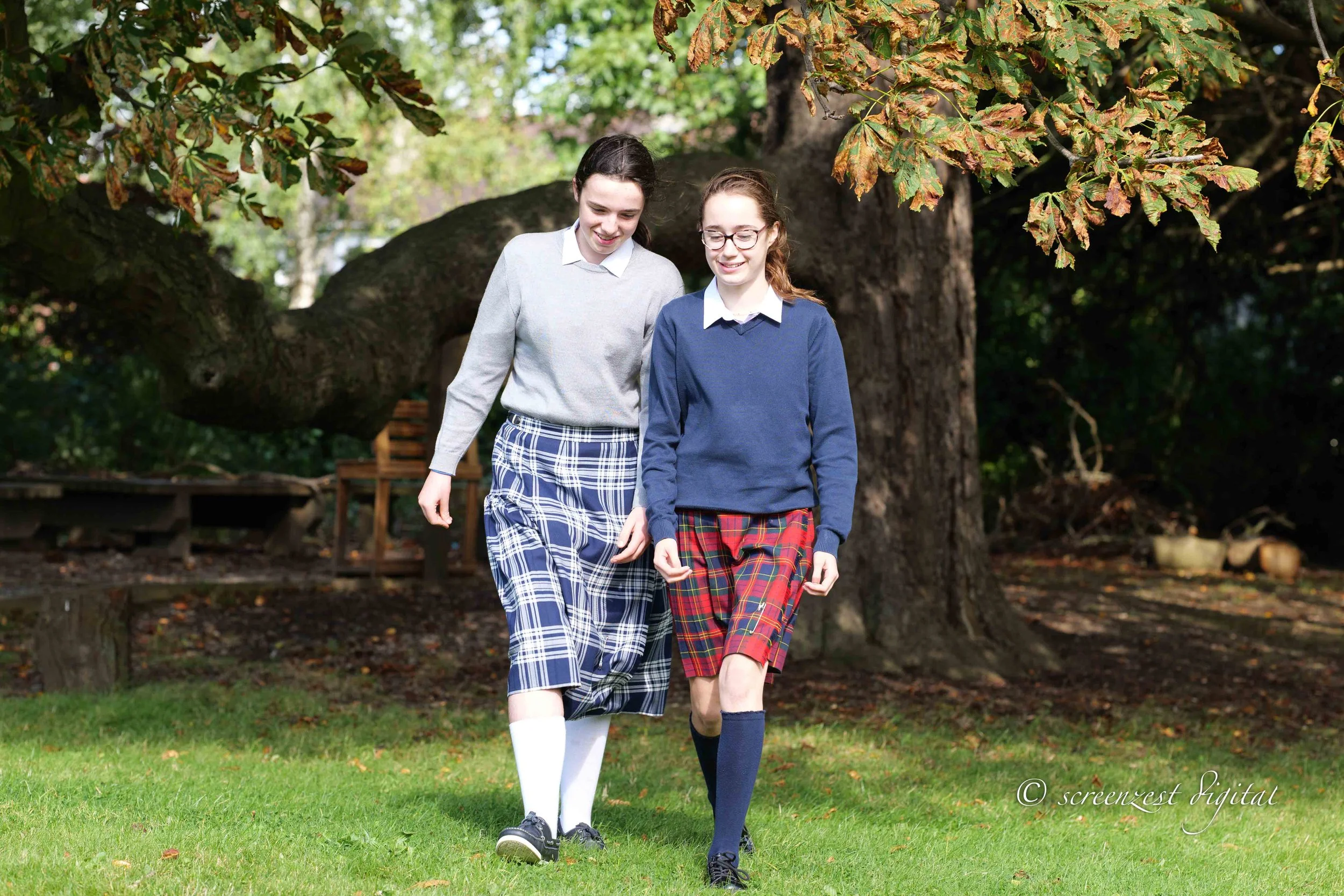 Two young women walking on grass in a park with large trees in the background, wearing school uniforms and plaid skirts.