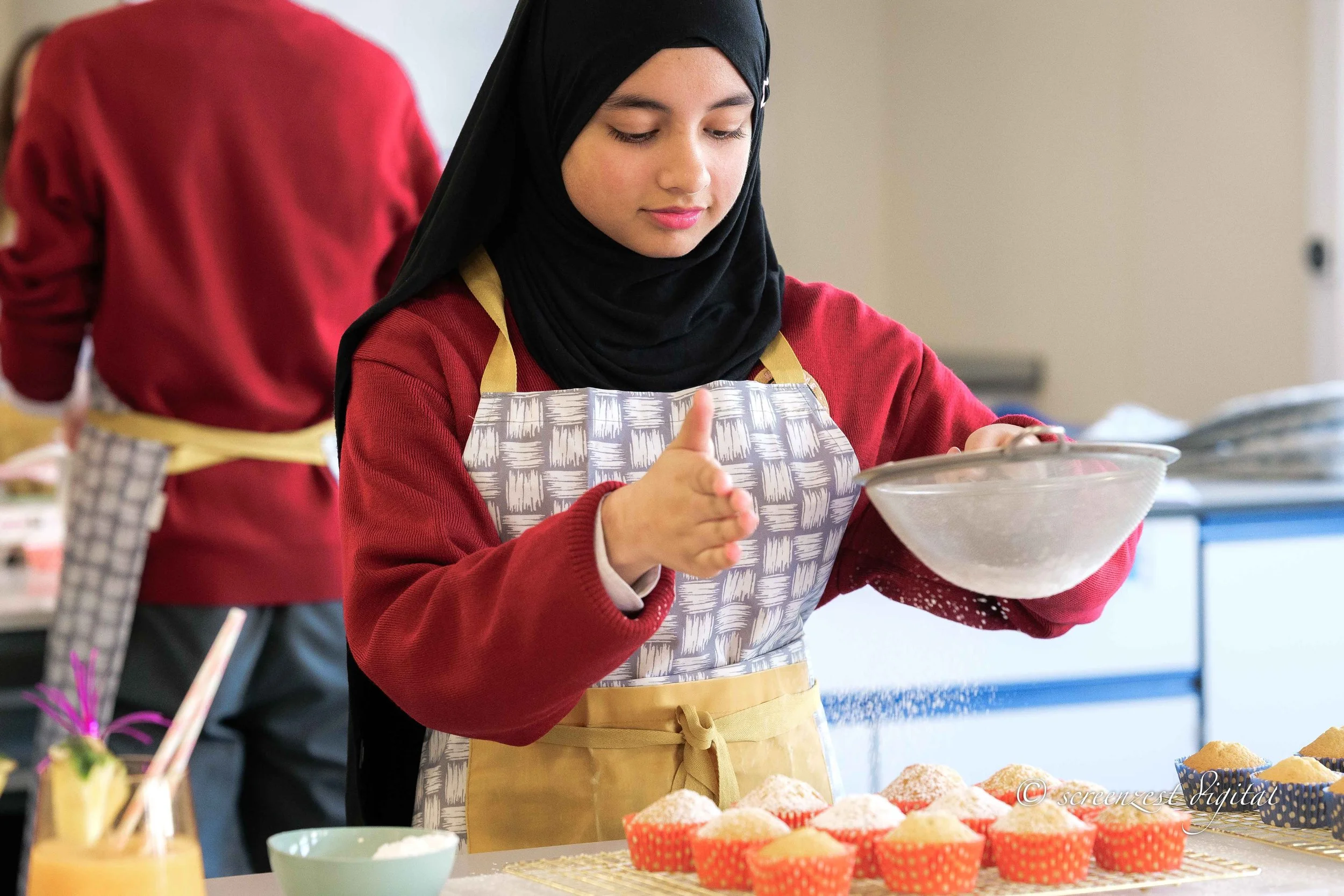 A young woman in a black hijab and red sweater with a beige apron is sifting flour into a mixing bowl while baking cupcakes in a kitchen. There are cupcakes in red and blue cupcake liners on the counter, along with other baking supplies, and a person