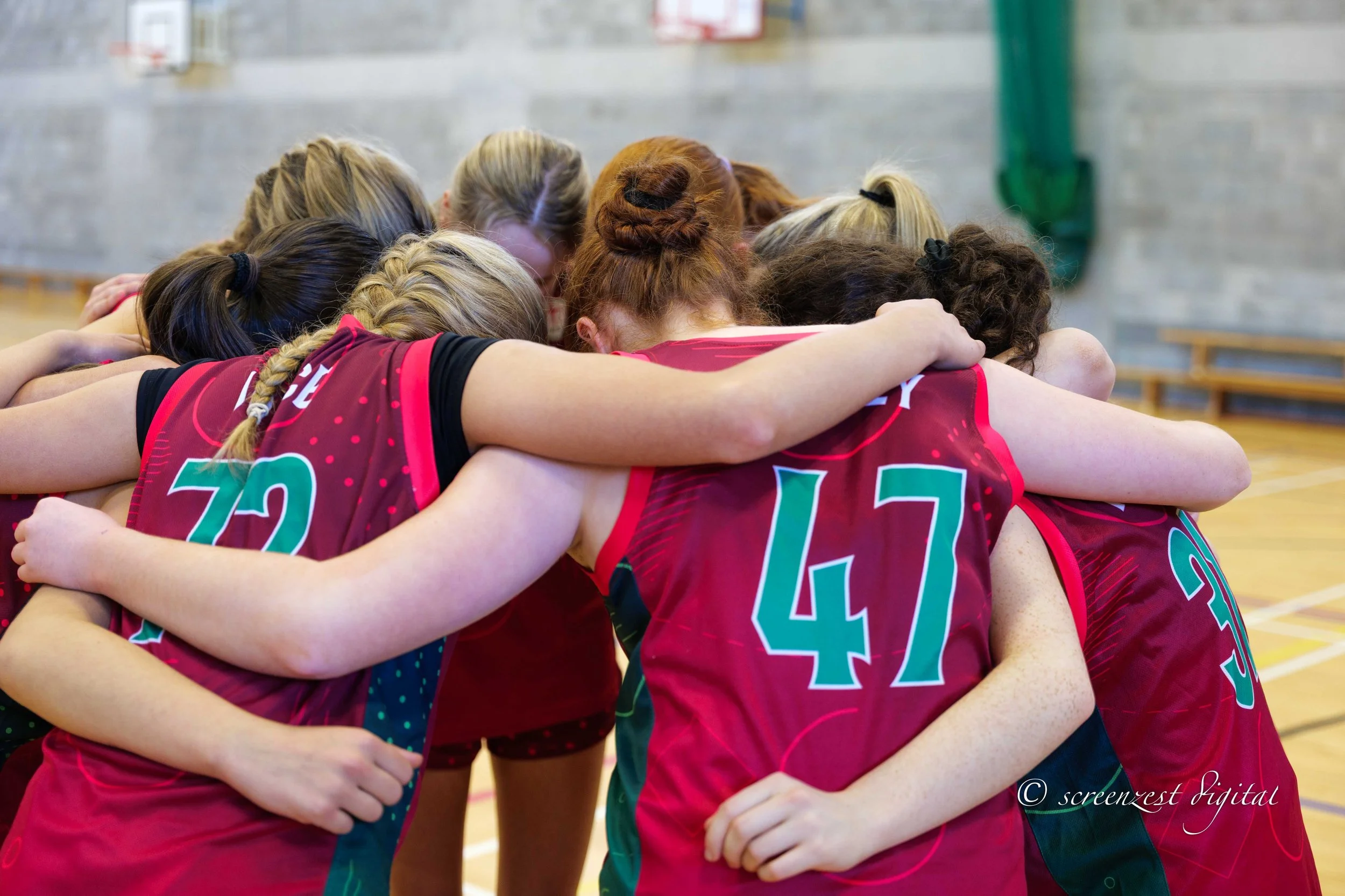 A group of female volleyball players wearing red jerseys with green numbers are in a huddle in a gymnasium, with arms around each other.