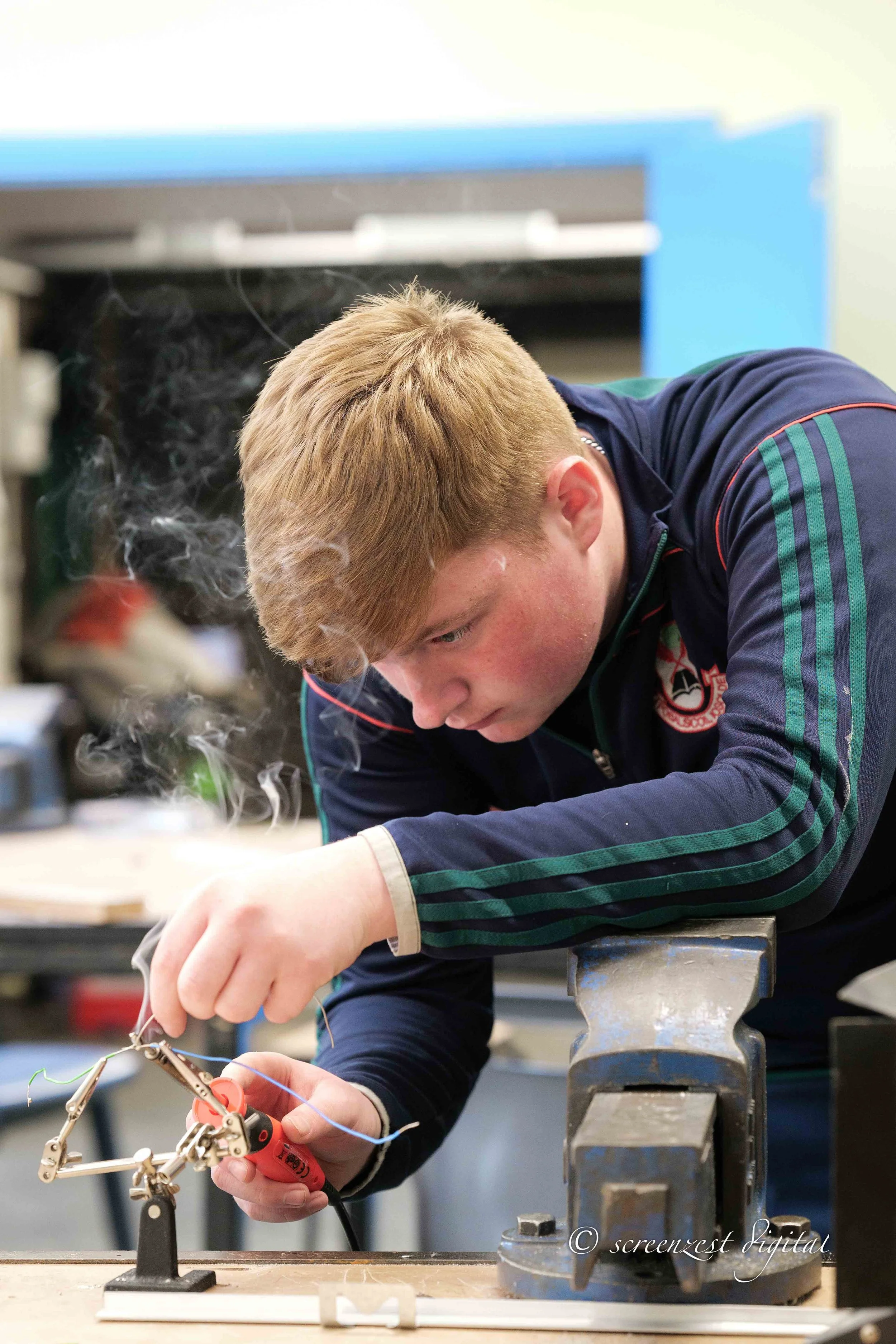 A young man with short, reddish-blonde hair working on a mechanical project at a workbench. He's wearing a navy blue jacket with green stripes on the sleeves.