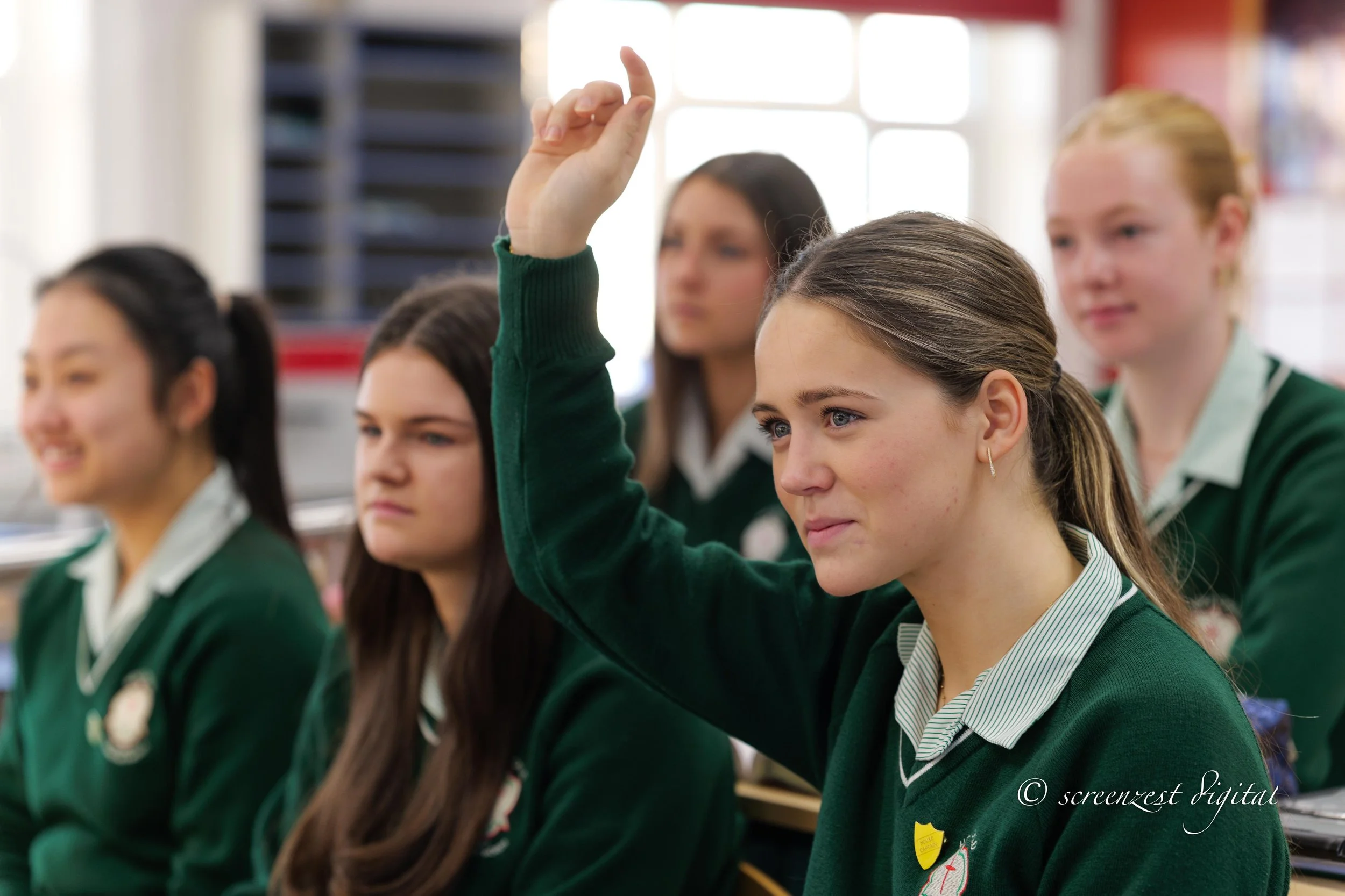 A classroom with five female students in green uniforms. One student, in the foreground, raises her hand, while the others sit and look ahead.