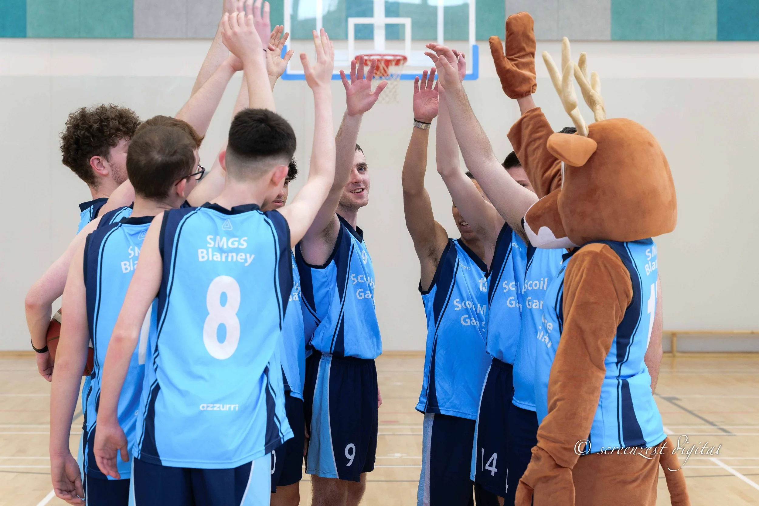 A group of young male basketball players in blue jerseys giving each other high fives on a basketball court, with a person in a reindeer costume celebrating with them.