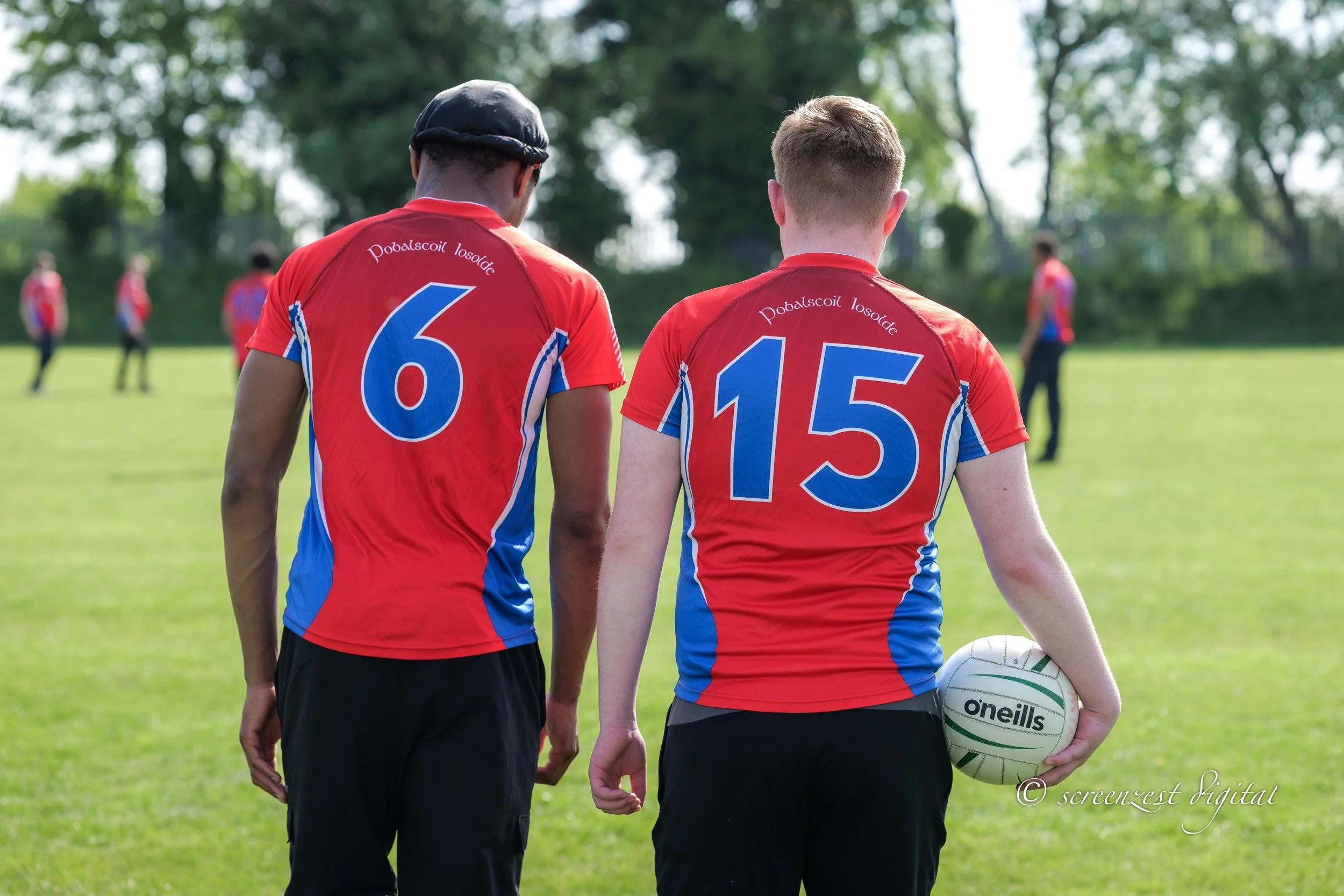 Two rugby players walking on a field, one holding a rugby ball, with a few more players in the background.