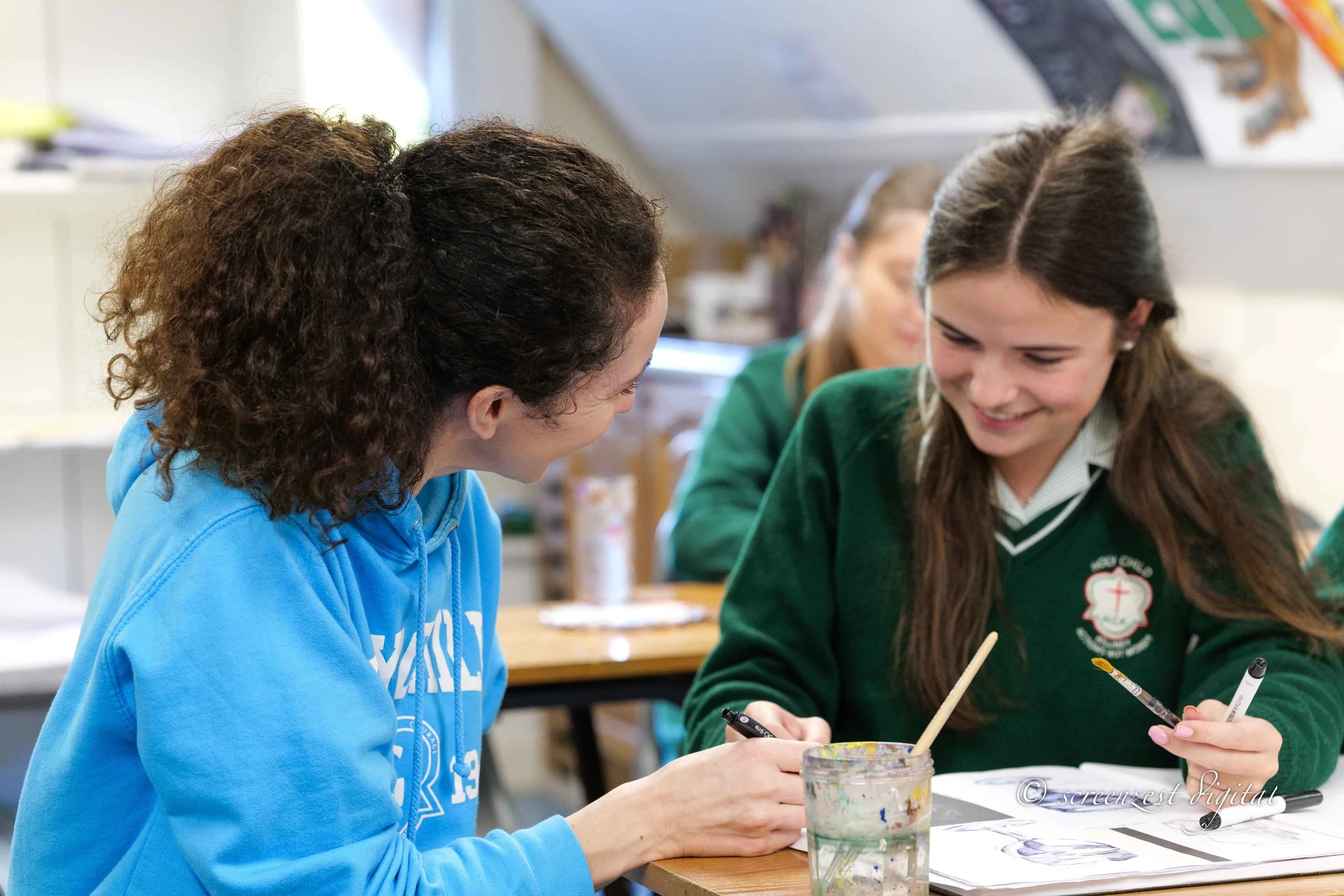 Two girls sitting at a table, smiling and engaging in an art activity with paintbrushes and papers, in a classroom setting.
