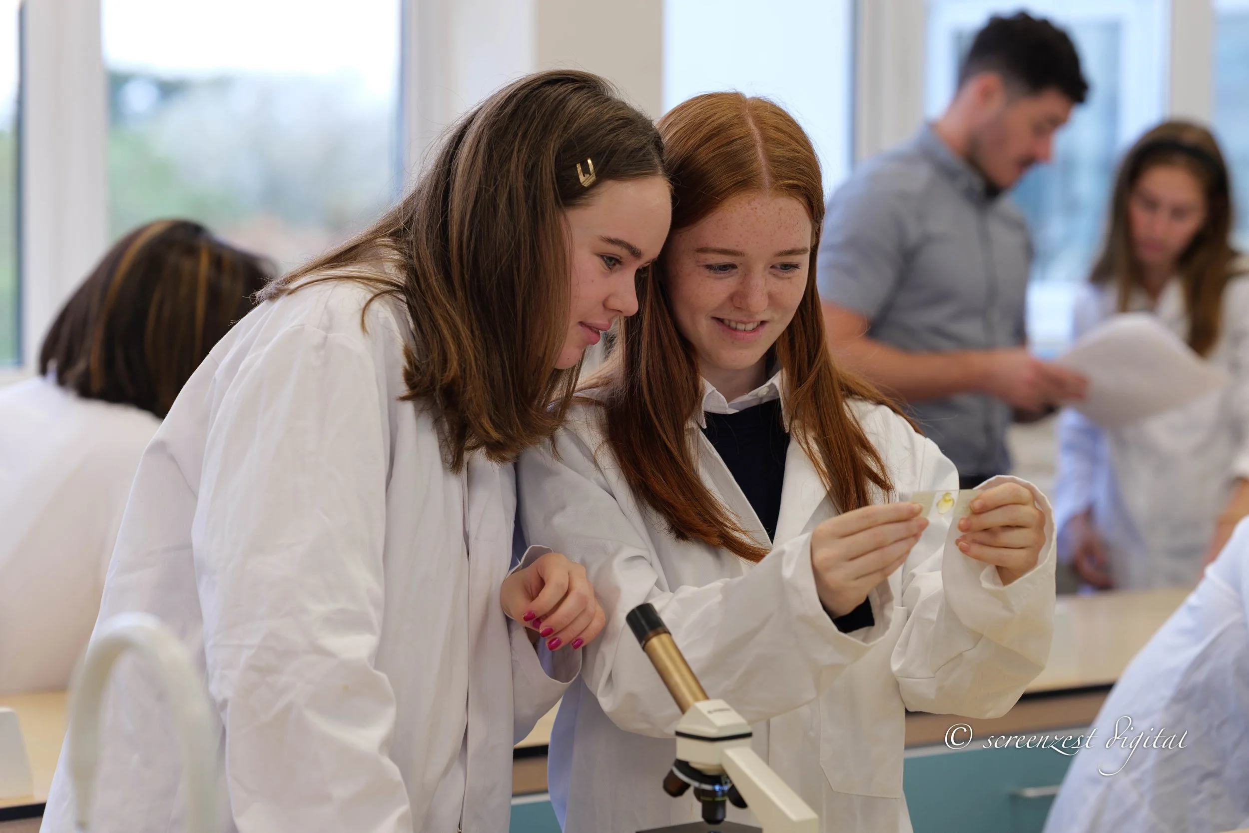 Two schoolgirls in white lab coats examining a small specimen in a classroom with other students and a microscope nearby.