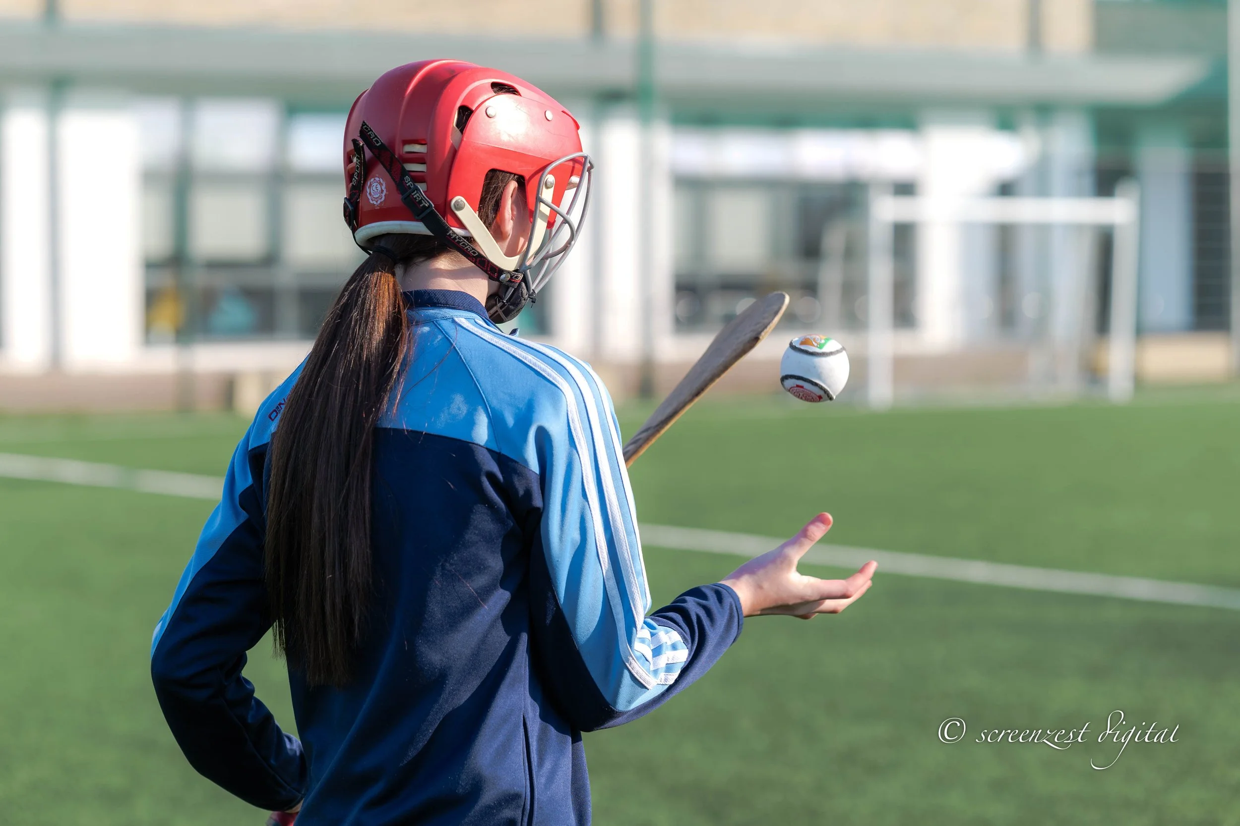 A young woman wearing a blue sports jacket and a red helmet juggles a white ball with colored details on a grassy field.