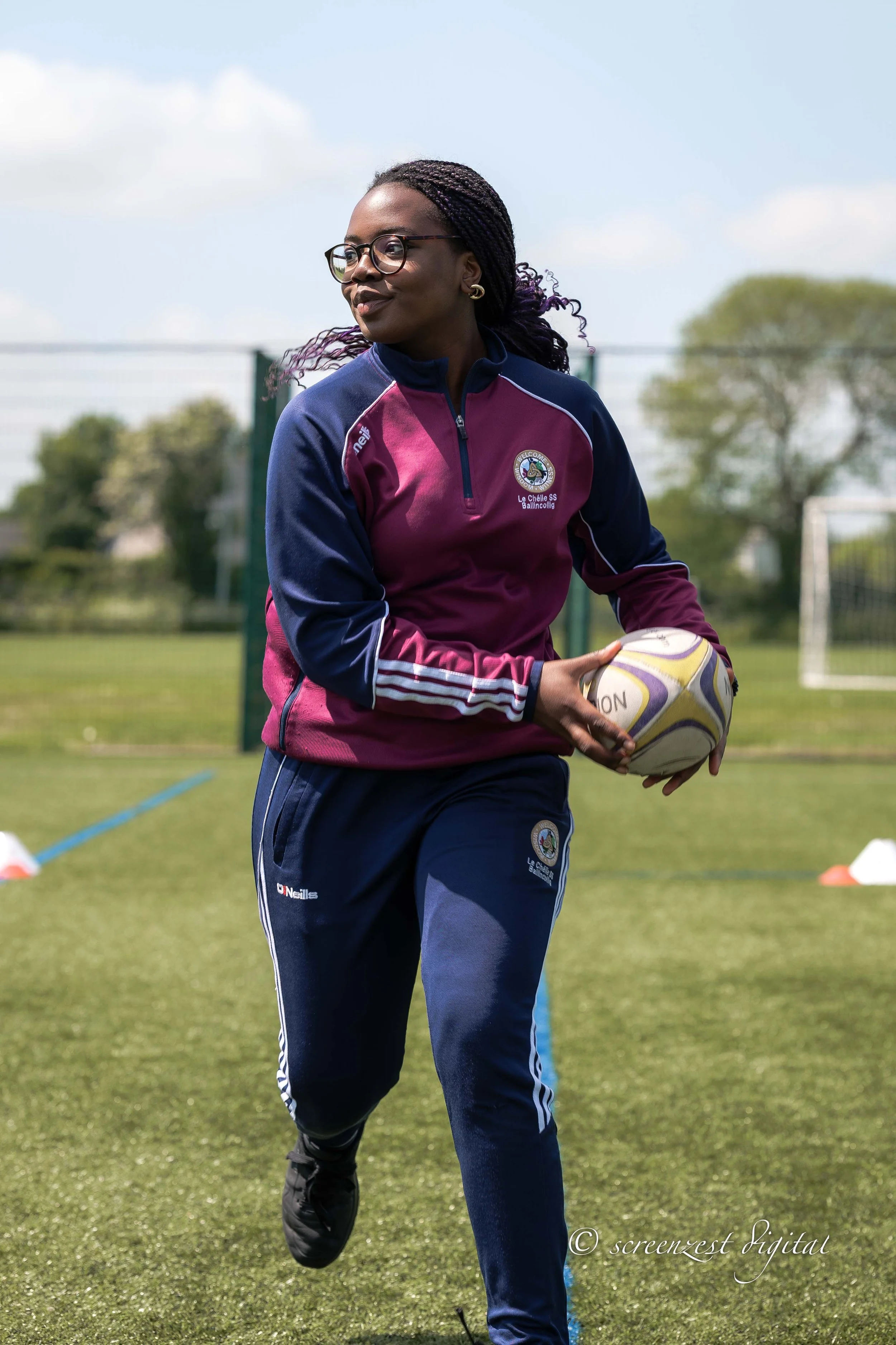 Young woman holding a rugby ball on a sports field wearing a maroon and navy sports jacket and navy sports pants with glasses and braided hair.