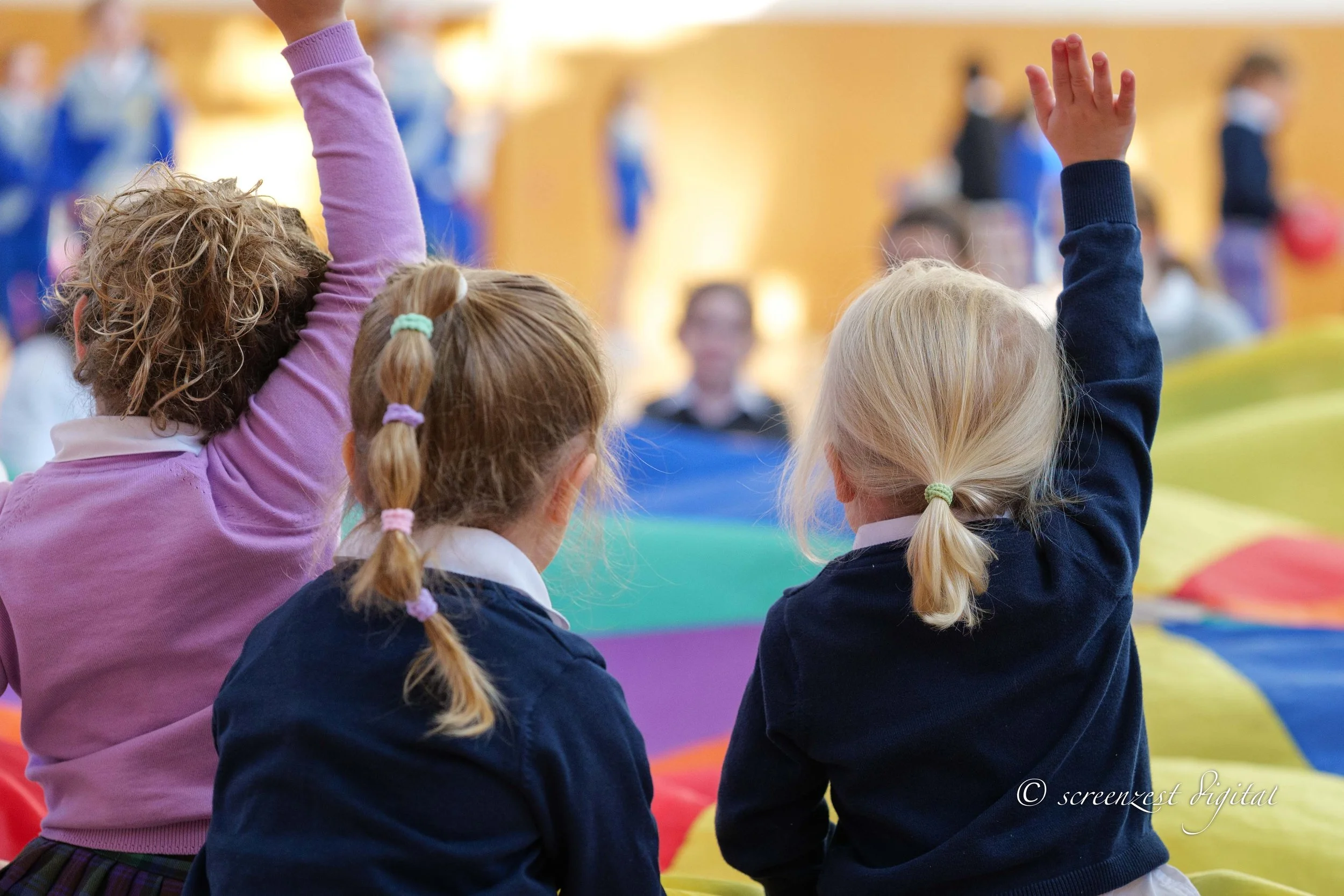 Three young girls in school uniforms sitting in a classroom or assembly hall. Two girls have raised their hands, and the third girl is listening. They are facing forward, and other children are visible in the background.