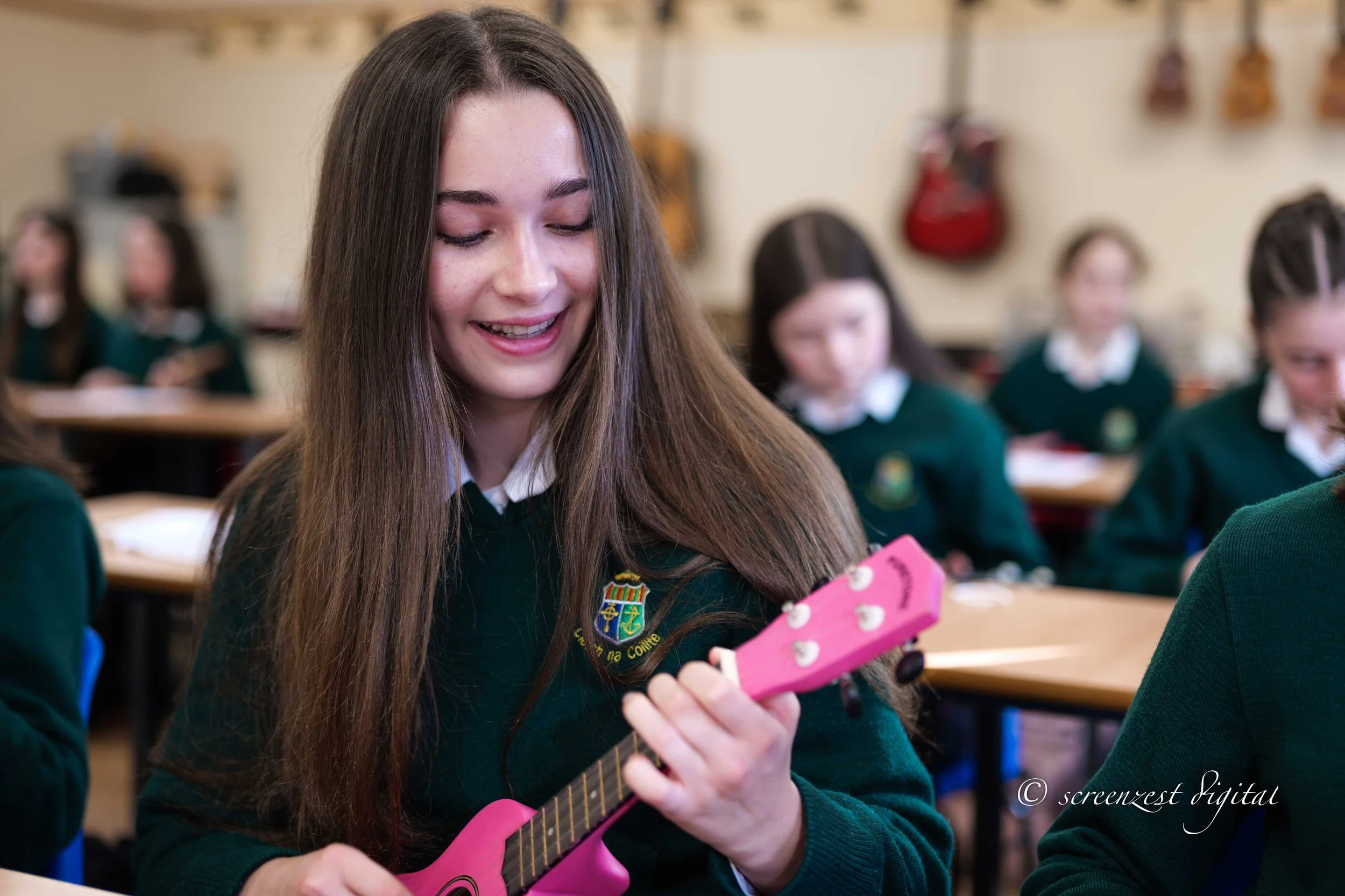A smiling teenage girl with long brown hair plays a small pink ukulele in a classroom. She is surrounded by other girls in green school uniforms, sitting at desks, with musical instruments hanging on the wall behind them.