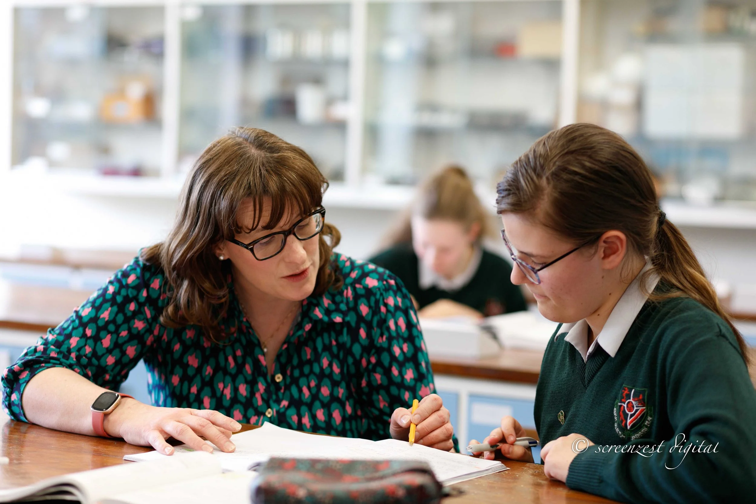 A teacher and a student working together at a classroom table with books and writing materials, engaged in a focused discussion.