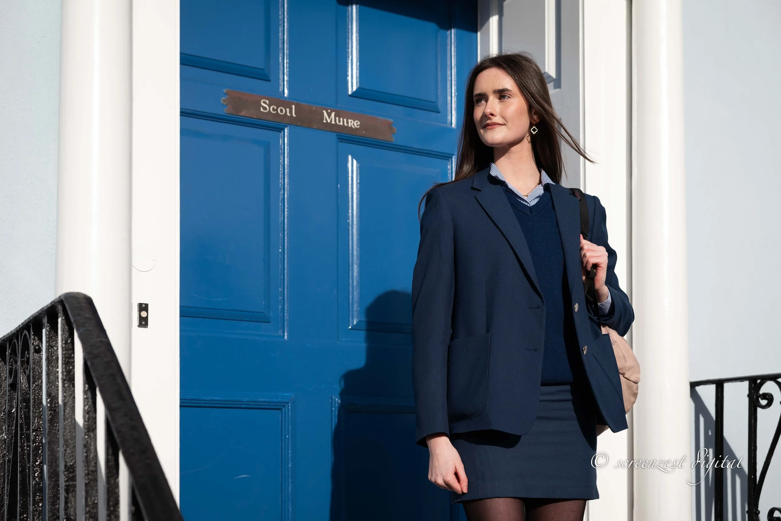 A young woman in a navy business suit and skirt standing in front of a blue door with the sign 'Scol Muire' on it, holding a beige bag on her shoulder.