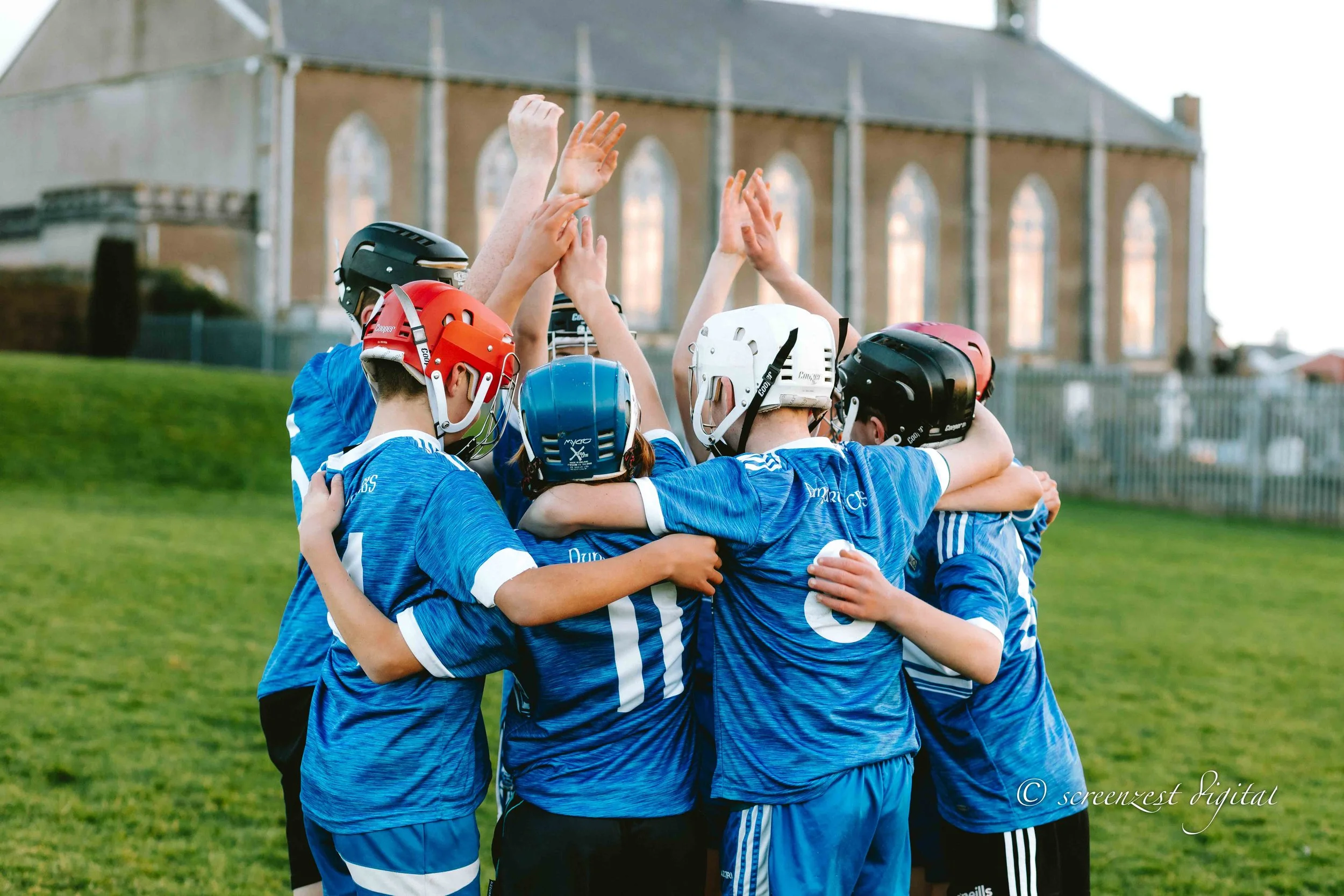 A group of young kids in blue sports uniforms and helmets are huddled together in a circle, celebrating on a grassy field with a church or large building in the background.