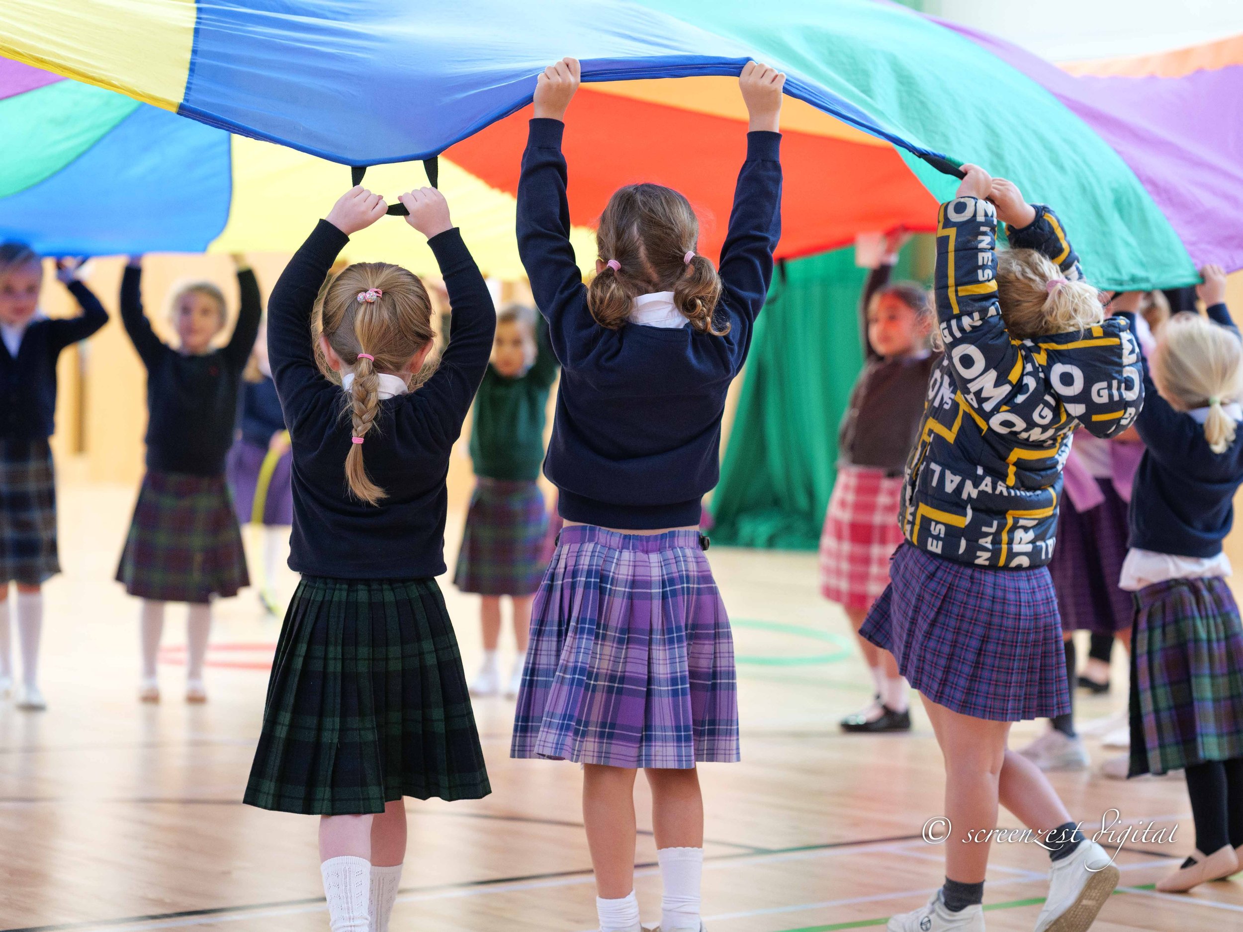 Children in school uniforms participating in a parachute game in a gymnasium.