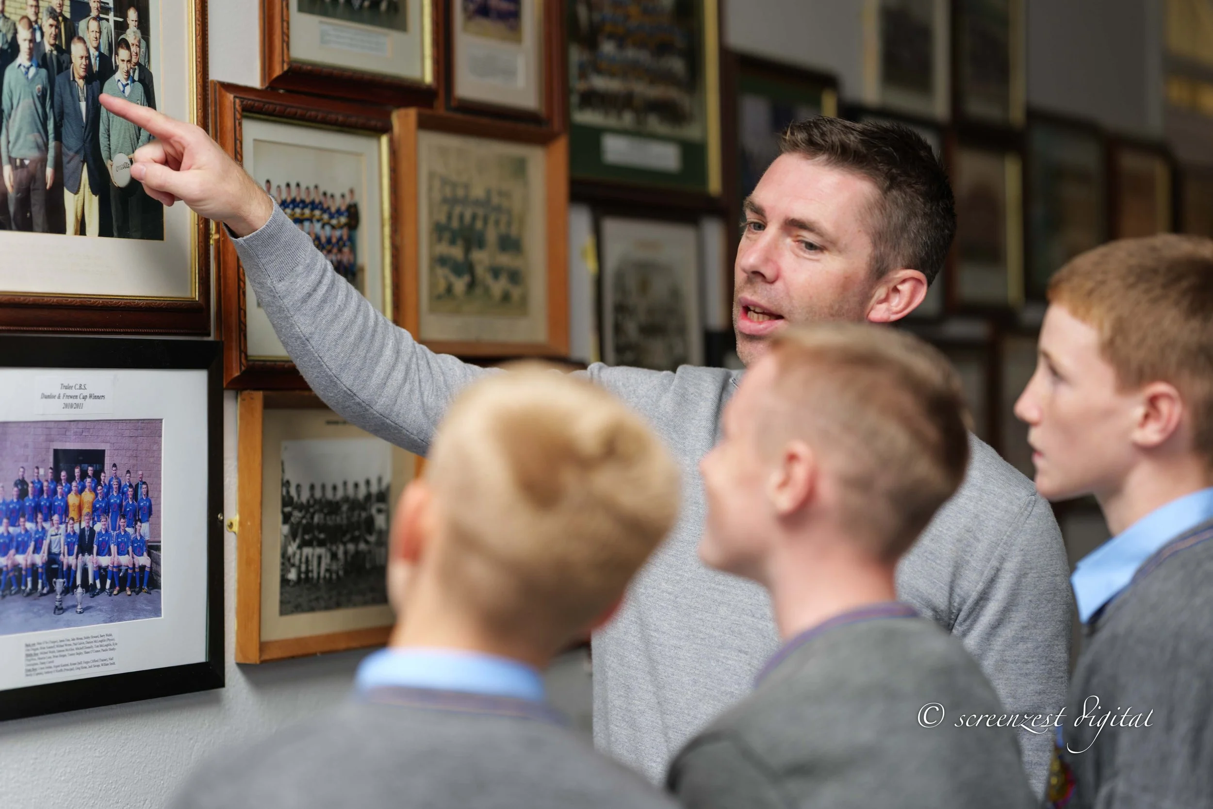 A man in a gray sweater is pointing at a framed photograph of a sports team on the wall, while three boys in school uniforms look on.