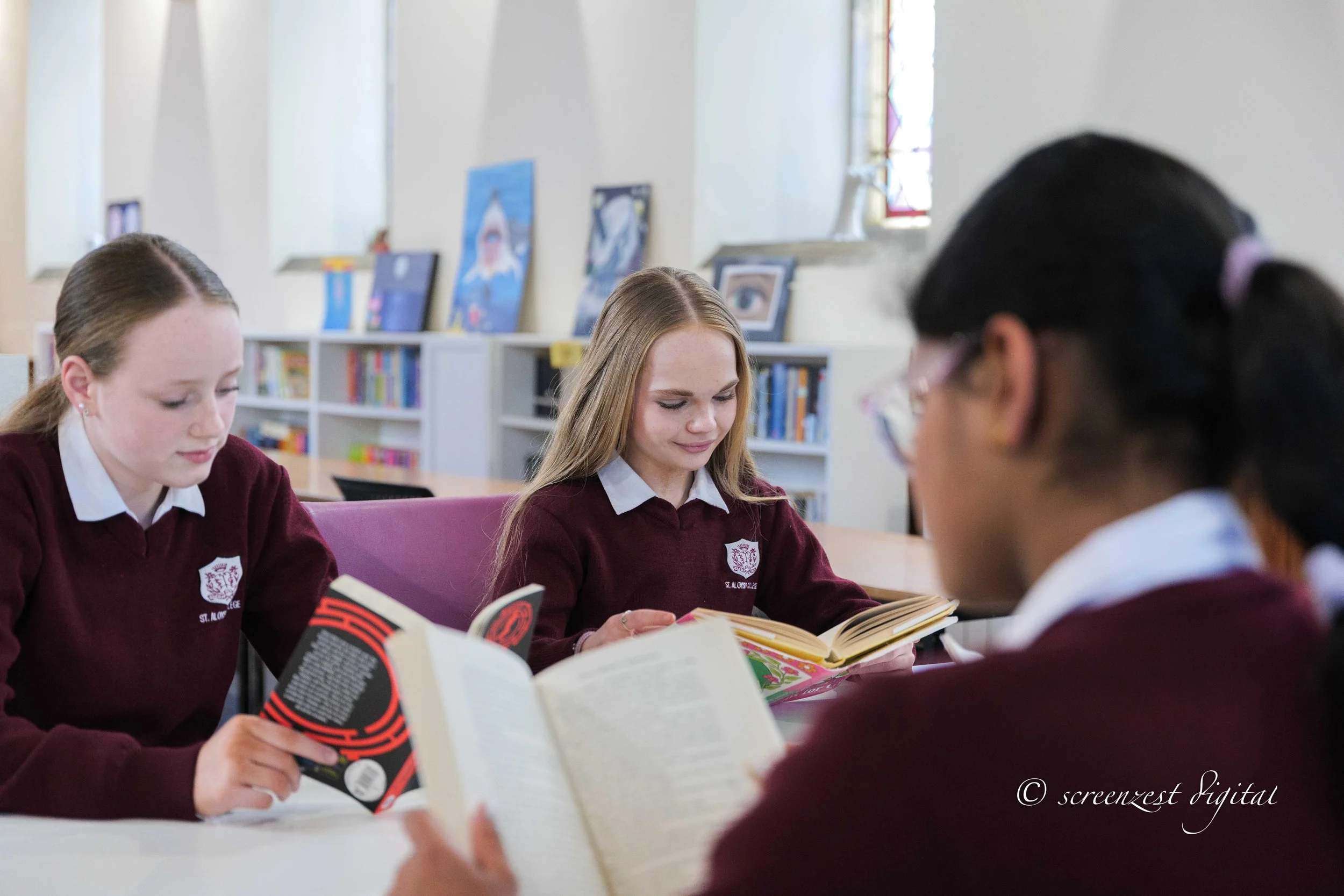 Three female students in school uniforms reading books at a table in a library or classroom.