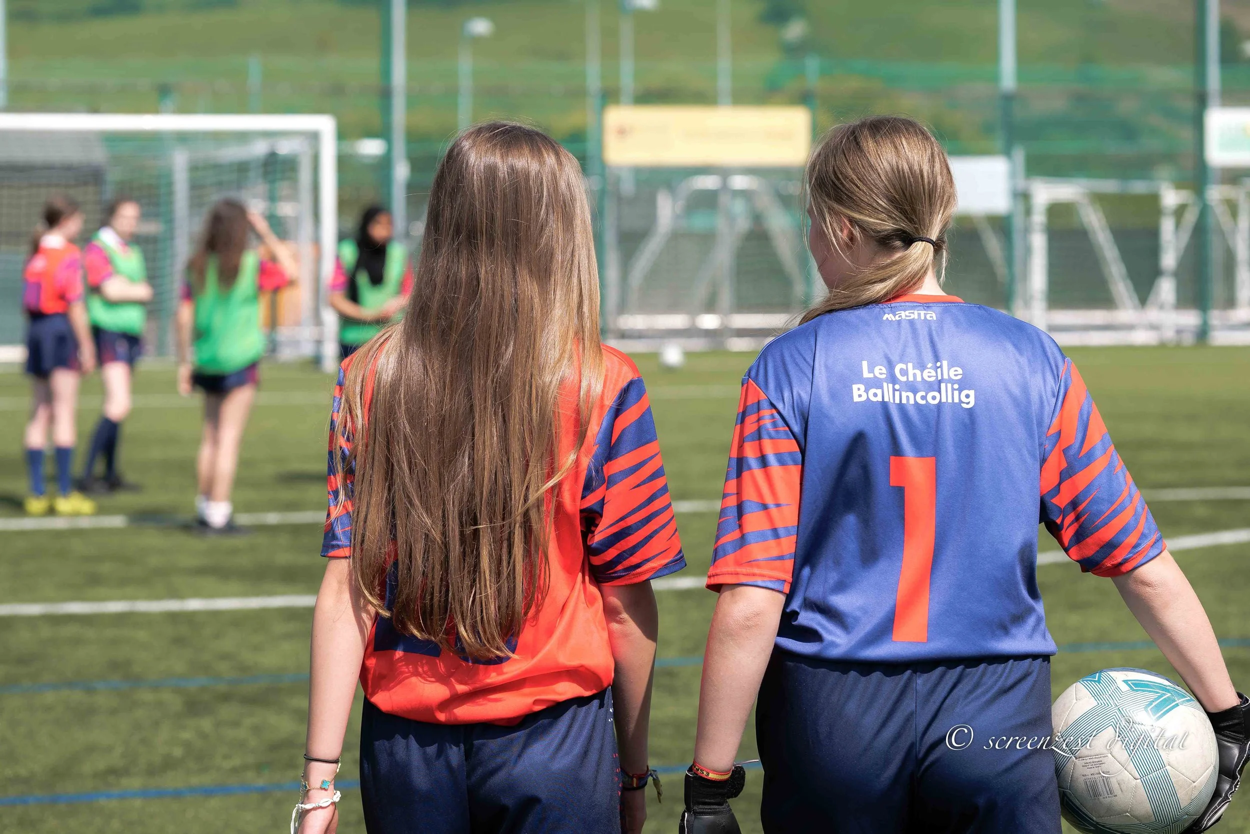 Two young girls in soccer uniforms stand on a soccer field with their backs to the camera, watching a group of girls in the distance. One girl has long brown hair and is wearing an orange and blue jersey. The other girl has blonde hair tied back and 
