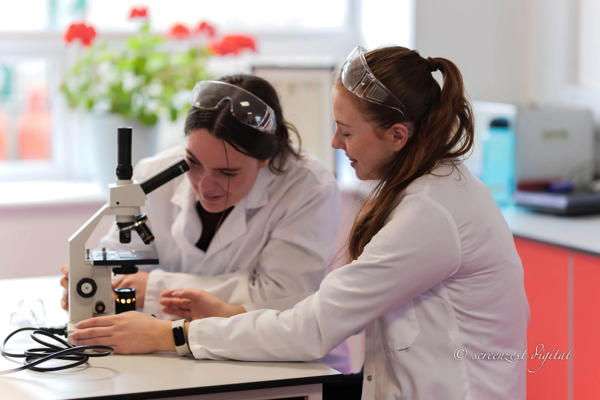 Two female scientists in lab coats and safety goggles working together with a microscope in a laboratory. One is looking through the microscope, the other is pointing at the slide, both smiling.