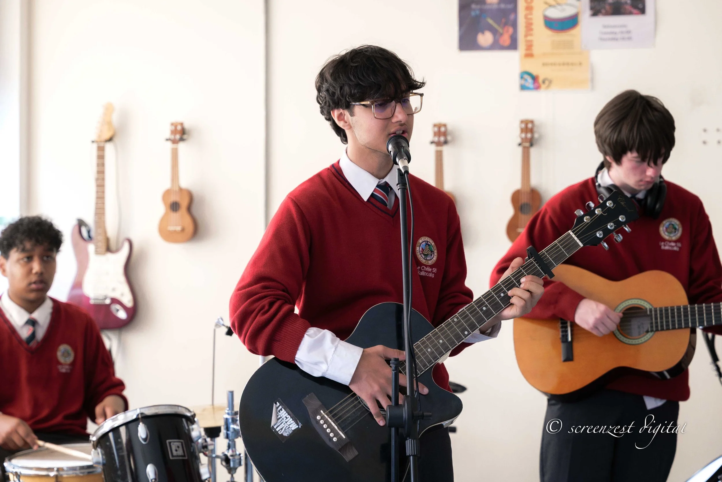 Three students in red school uniforms playing guitars and drums during a musical performance in a classroom with ukulele wall decorations.