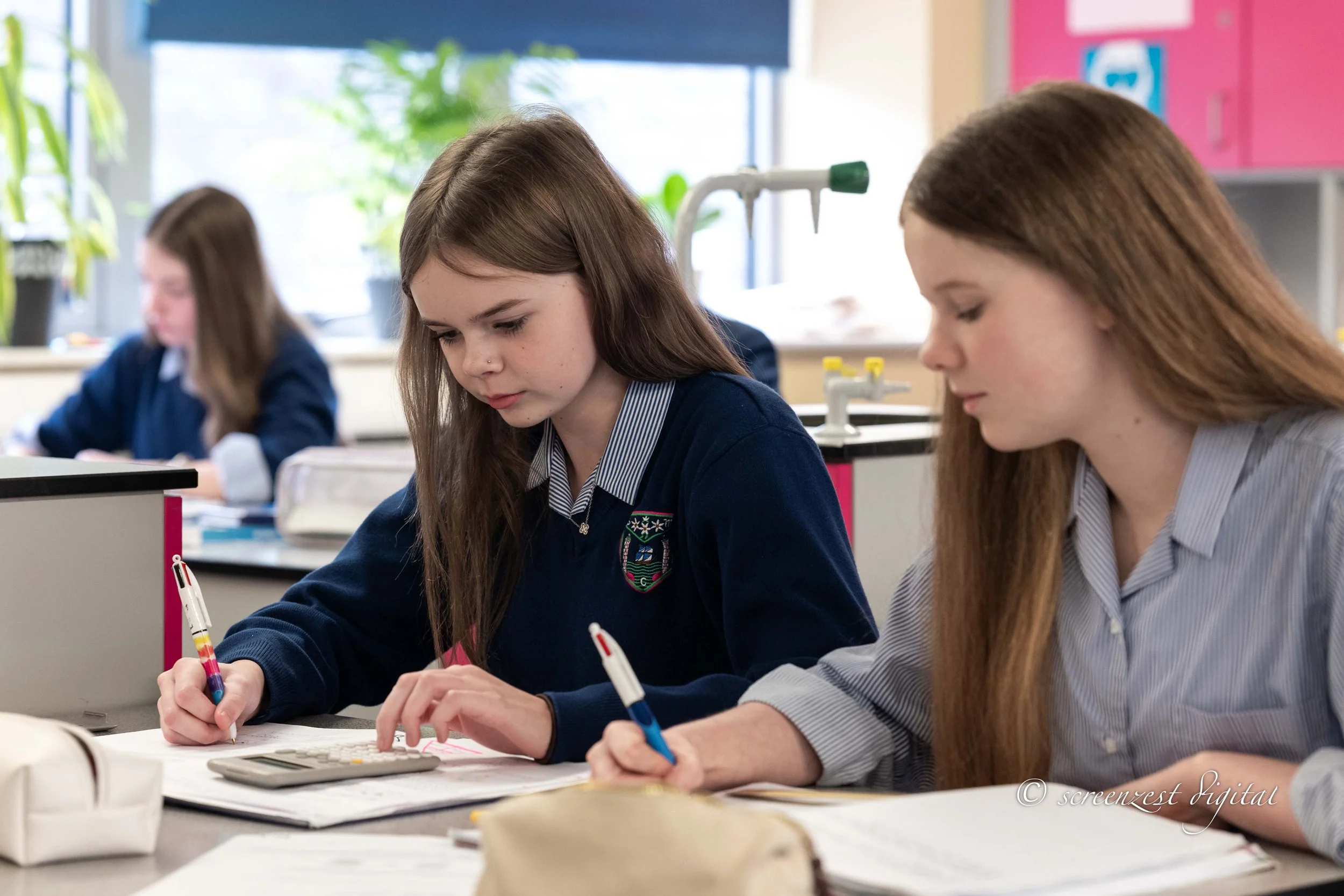Two female students working on a math assignment at a desk in a classroom, with a calculator and papers, while studying.