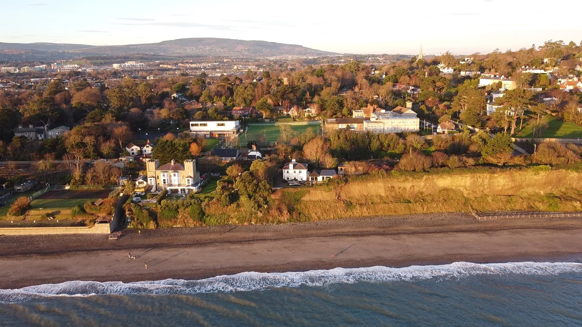 Aerial view of houses, trees, and a football field along a beach shoreline with waves and hills in the background.