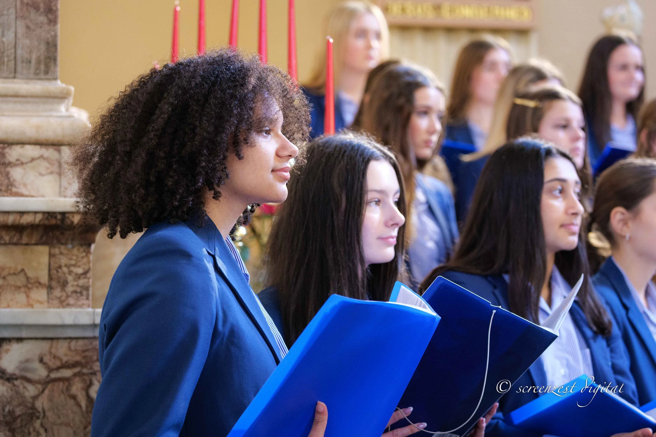A group of young women in blue robes, holding blue folders, participating in a formal ceremony or graduation in a church or similar venue with candles and decorative elements in the background.