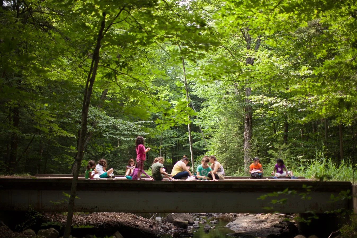 Young campers drawing in sketchbooks while sitting on bridge overlooking a creek surrounded by trees in the woods.