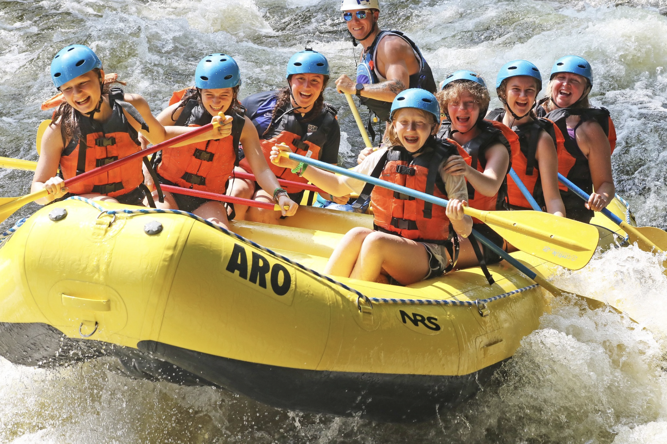 Happy campers smiling and laughing in a yellow raft while paddling through white water rapids.