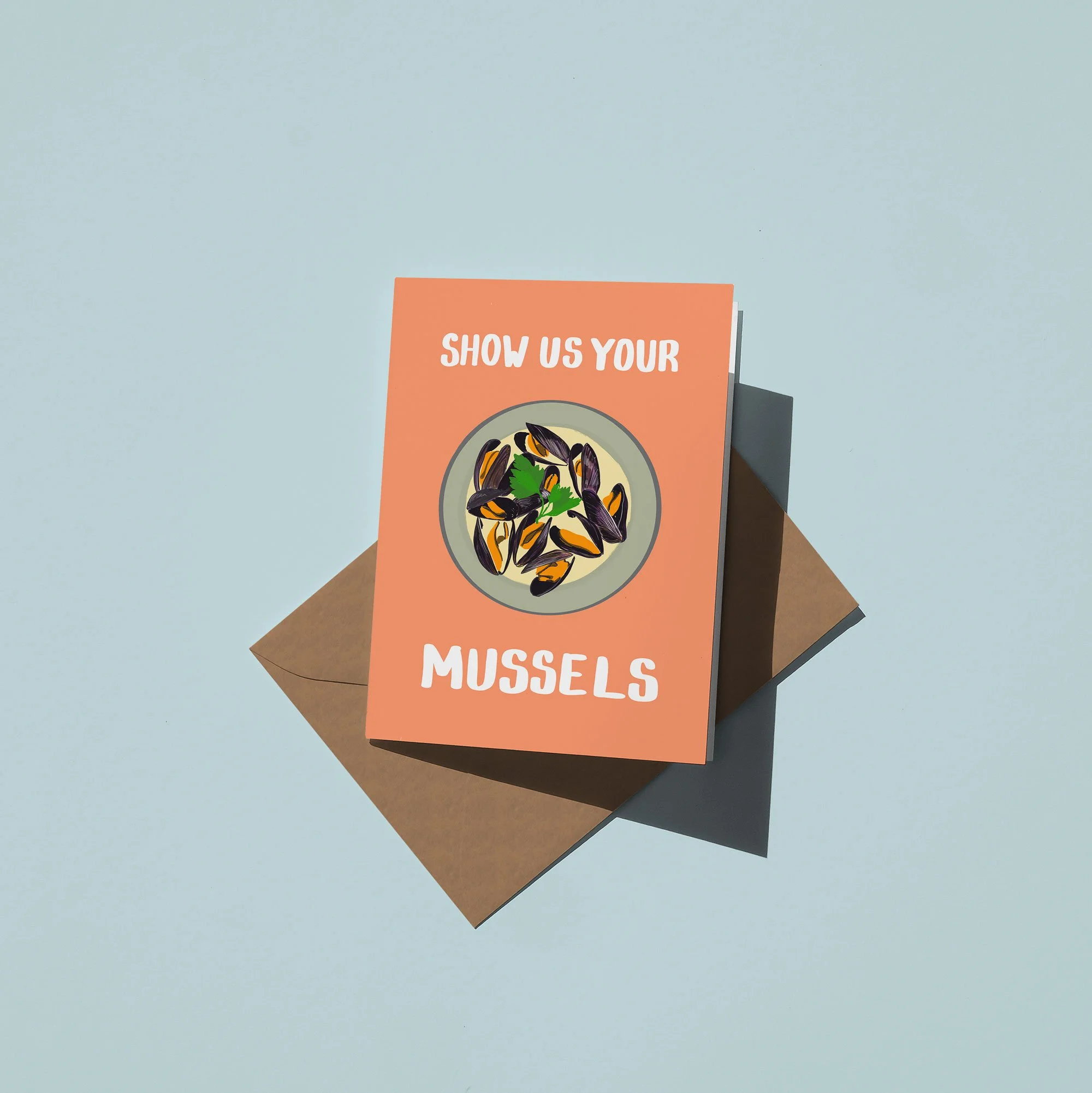 A greeting card with the message "Show us your mussels" and an illustration of mussels on a plate, placed on a brown envelope against a light background.