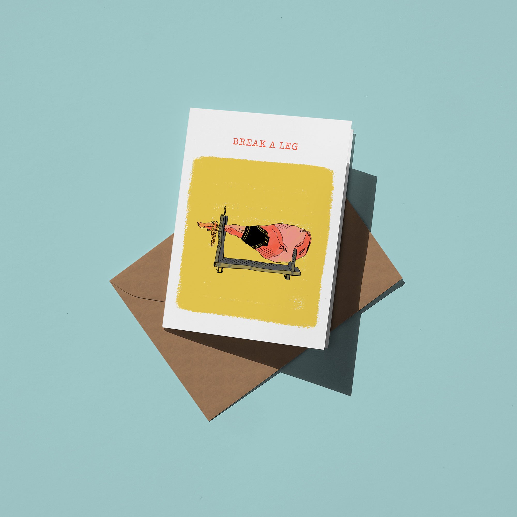 A greeting card with an illustration of a foot in black athletic wear resting on a wooden leg press machine, and the text 'Break a Leg' at the top, placed on top of an envelope on a blue surface.