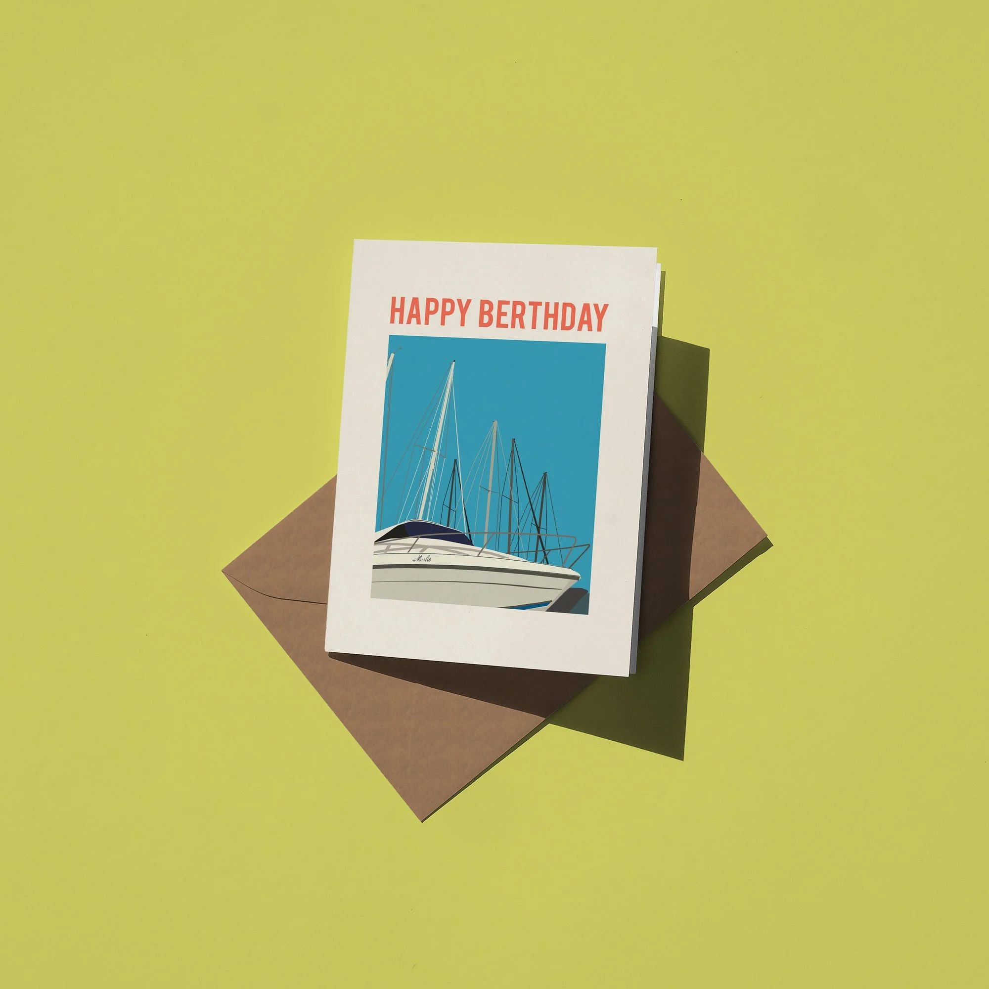 A birthday card with the message 'HAPPY BIRTHDAY' and an illustration of sailboats on the water, placed on a brown envelope on a yellow-green background.
