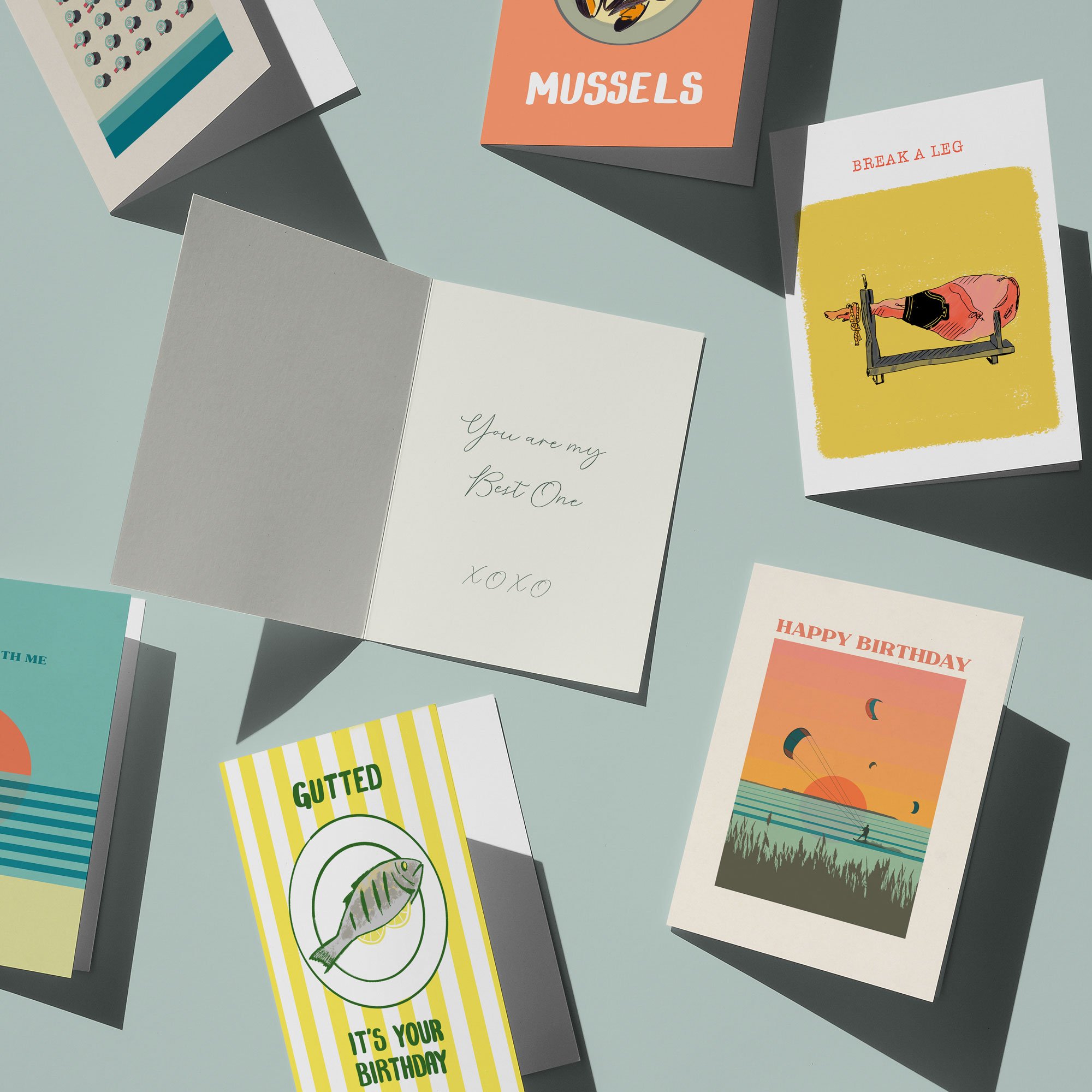 Mixed greeting cards on a light blue surface, including birthday, love, and humorous cards, with some cards open and others closed.