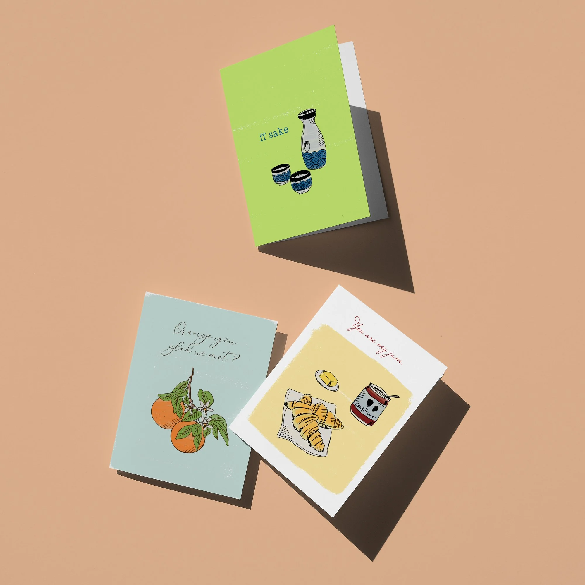 Three greeting cards on a beige background. The top card has a green background with an illustration of a sake set and the text 'if sake'. The bottom left card is light blue with an illustration of oranges on a branch and the text 'Orange you glad we