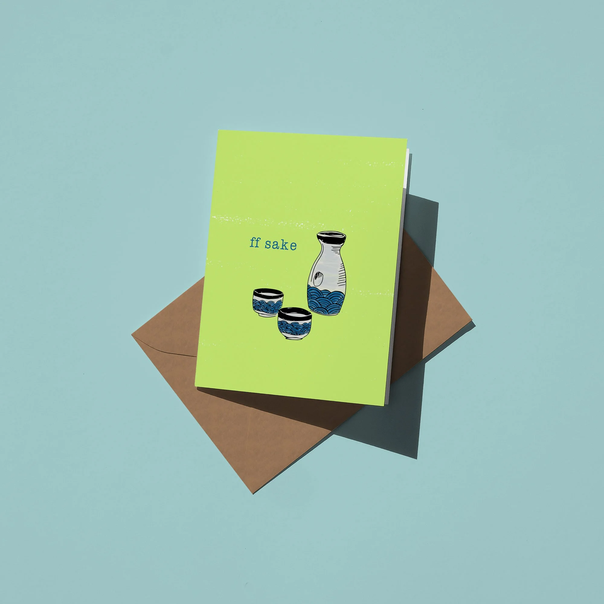 A green greeting card with blue and black illustrations of sake bottles and cups, placed on top of a brown envelope against a light blue background.