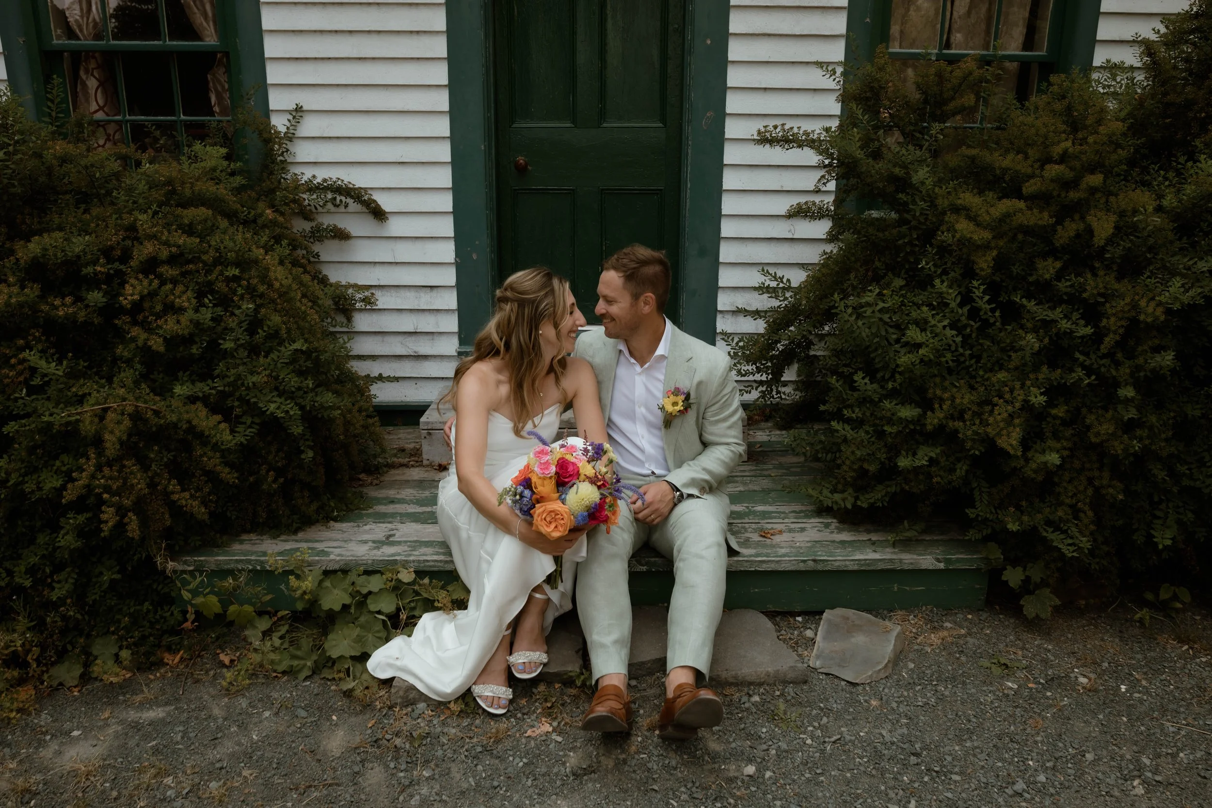 A couple, dressed in wedding attire, sitting on a rustic wooden porch in front of a white house with green trim. The woman holds a colorful bouquet of flowers and is smiling at the man, who is smiling back at her. Overgrown bushes surround them.