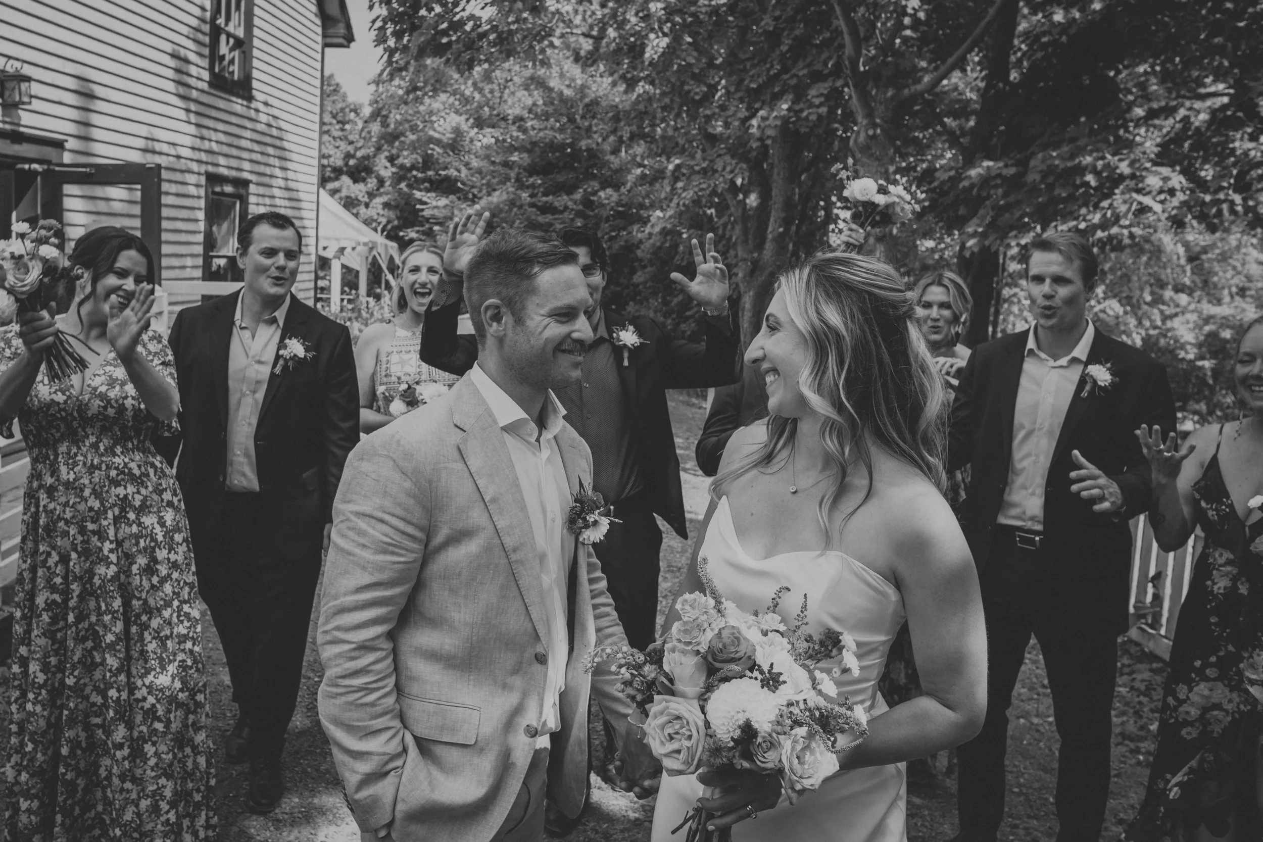 A black and white photo of a wedding ceremony outdoors. The bride and groom are smiling and facing each other, with the bride holding a bouquet. Guests are surrounding them, clapping and cheering.