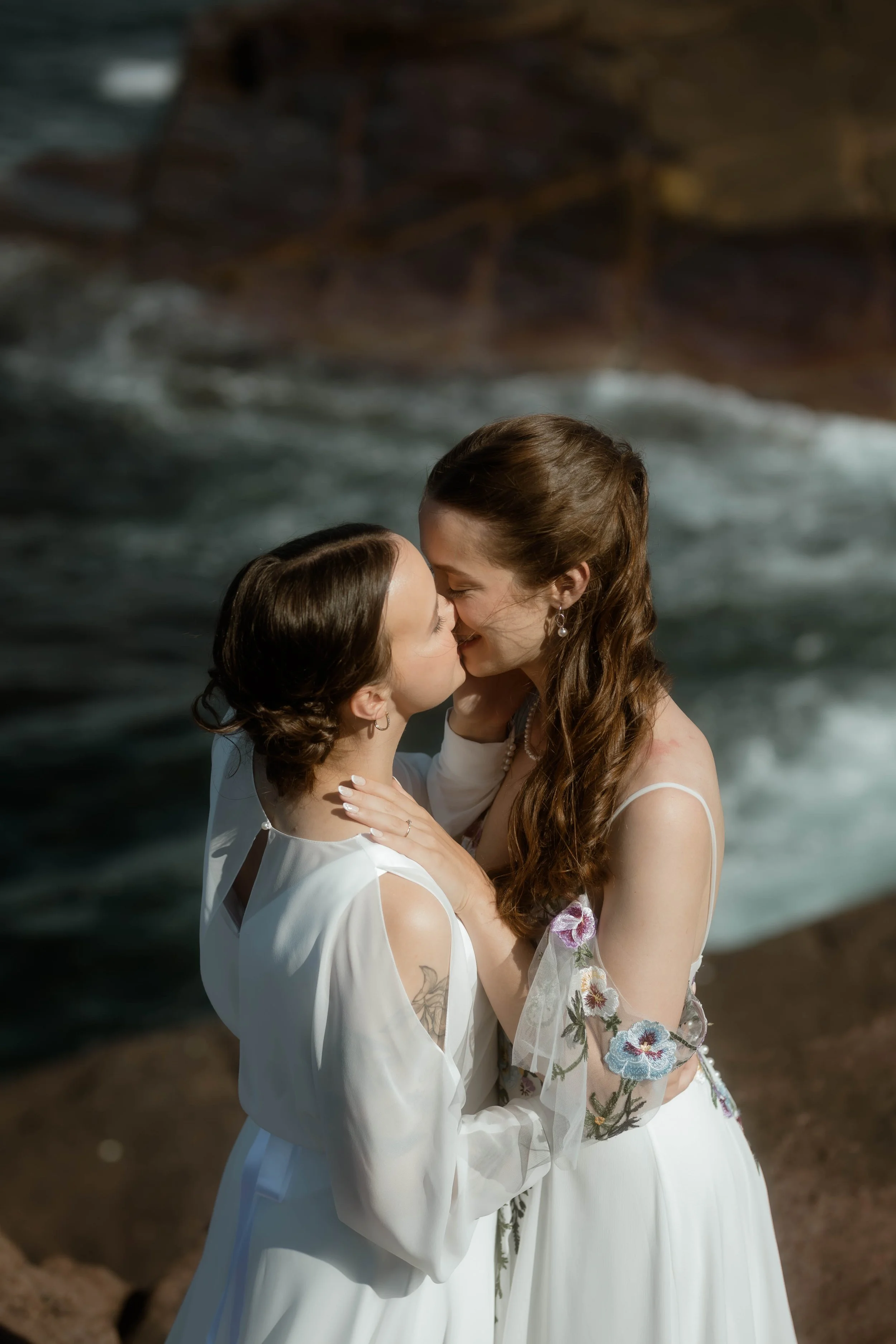 Two women in white dresses sharing a kiss on a beach with waves in the background.