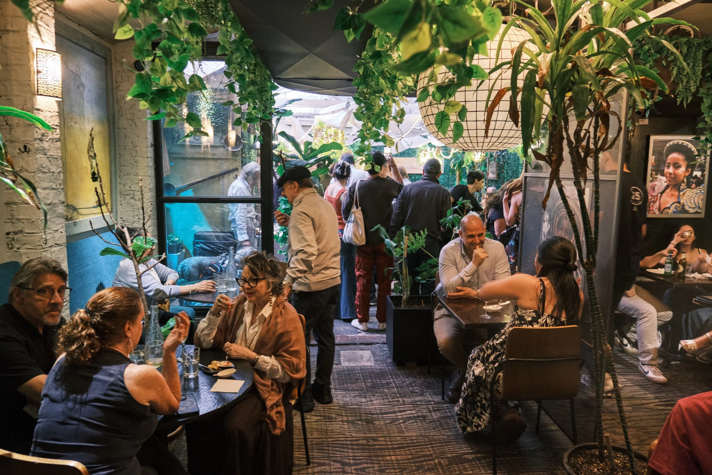 People dining and socializing in a cozy indoor restaurant with lush green plants, large windows, and artwork on the walls.