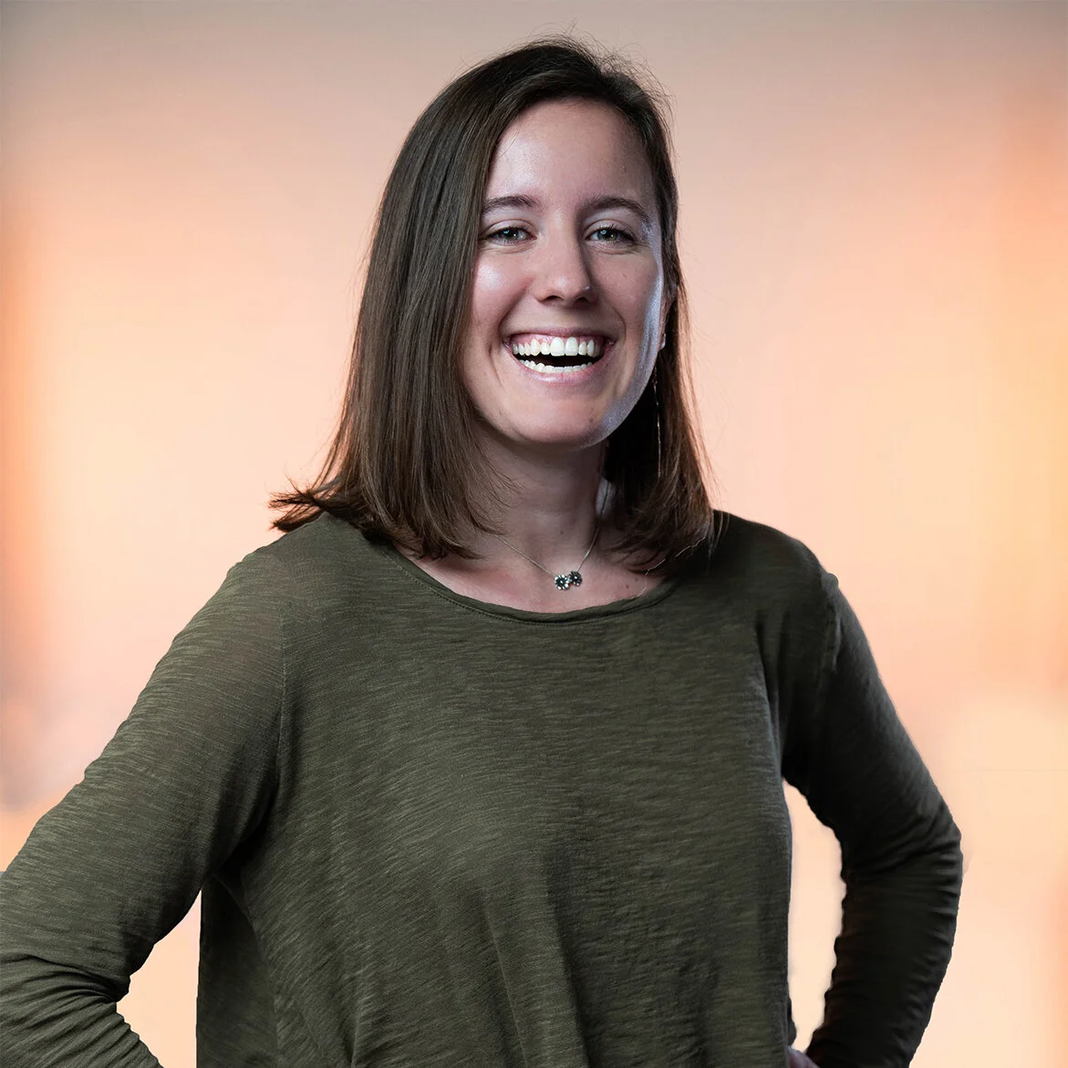 A white woman with shoulder length brown hair smiling and wearing a dark green long sleeve shirt standing against a light orange background